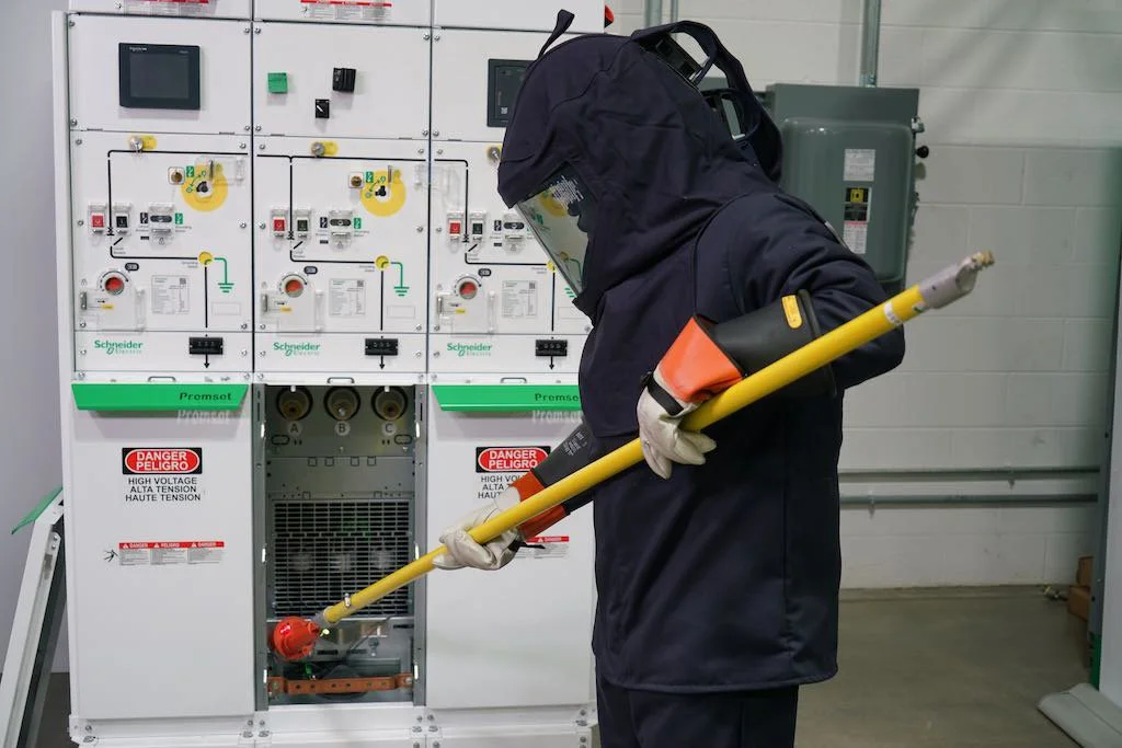 A worker in protective gear using a long-handled tool near electrical equipment in an industrial setting.