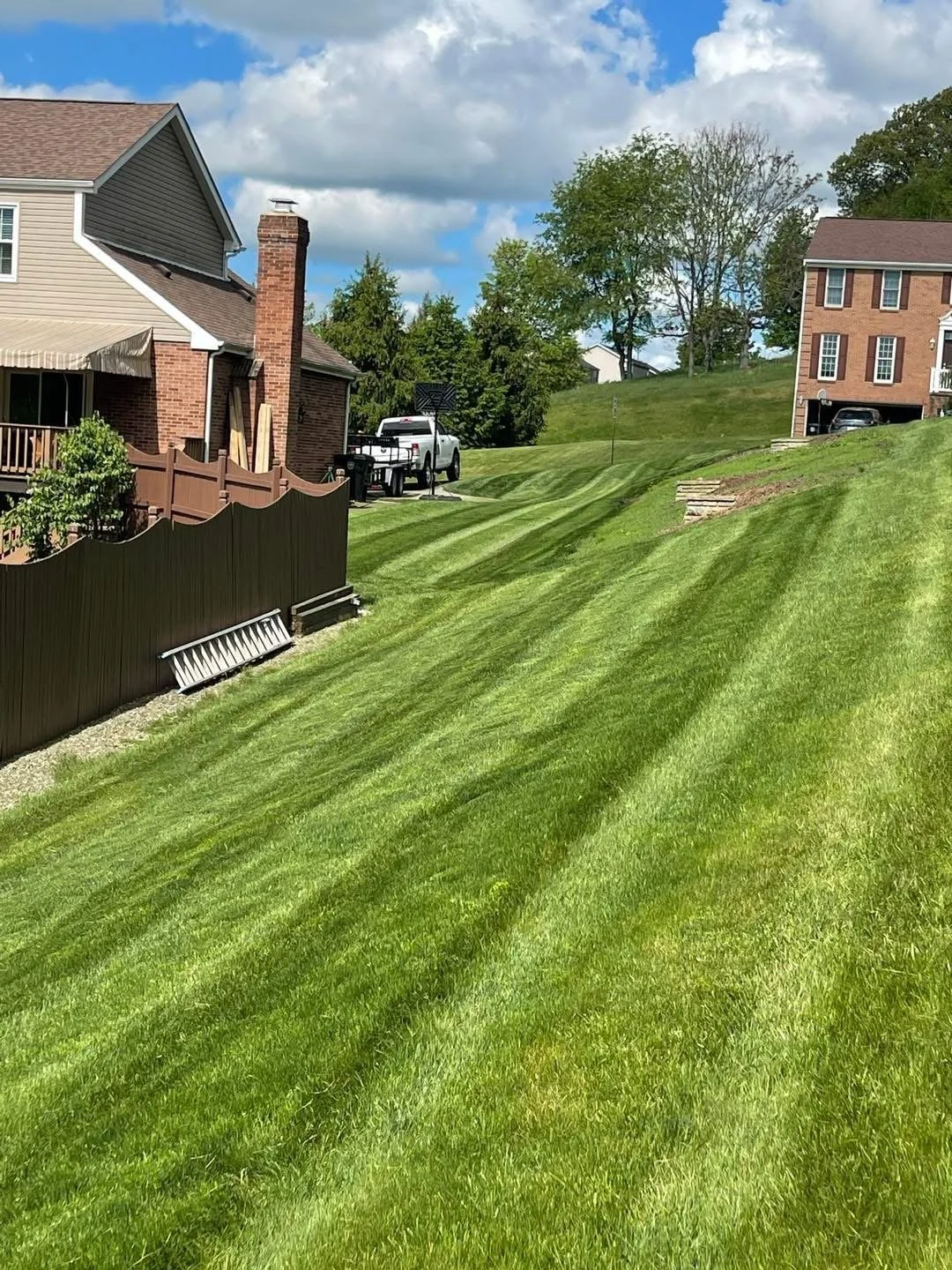 Well-maintained green backyard with striped mowing pattern, fenced area, partially visible brick and siding houses, and trees under a partly cloudy sky.