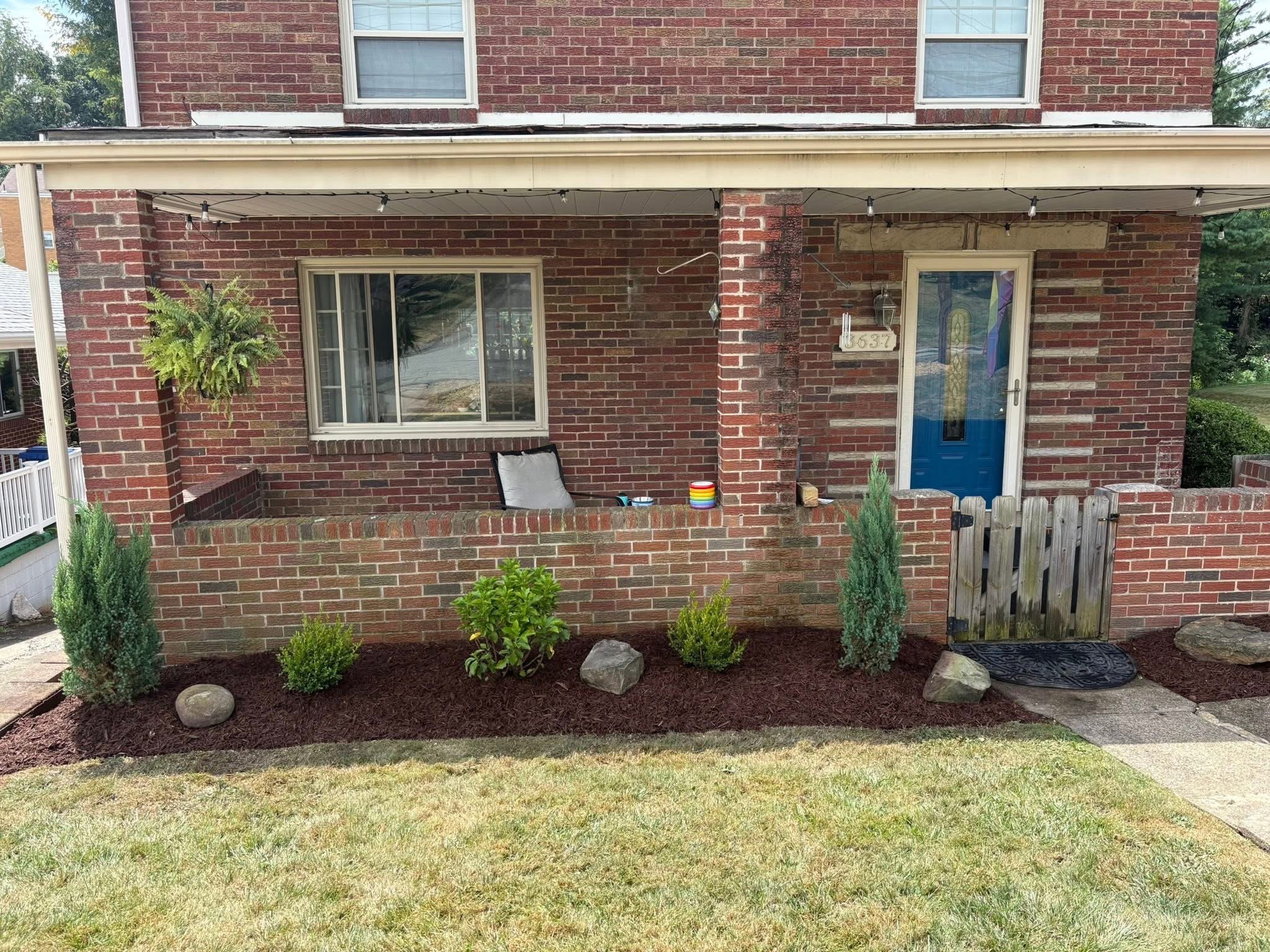 Front view of a brick house with a small porch, blue front door, and a garden with shrubs and rocks.