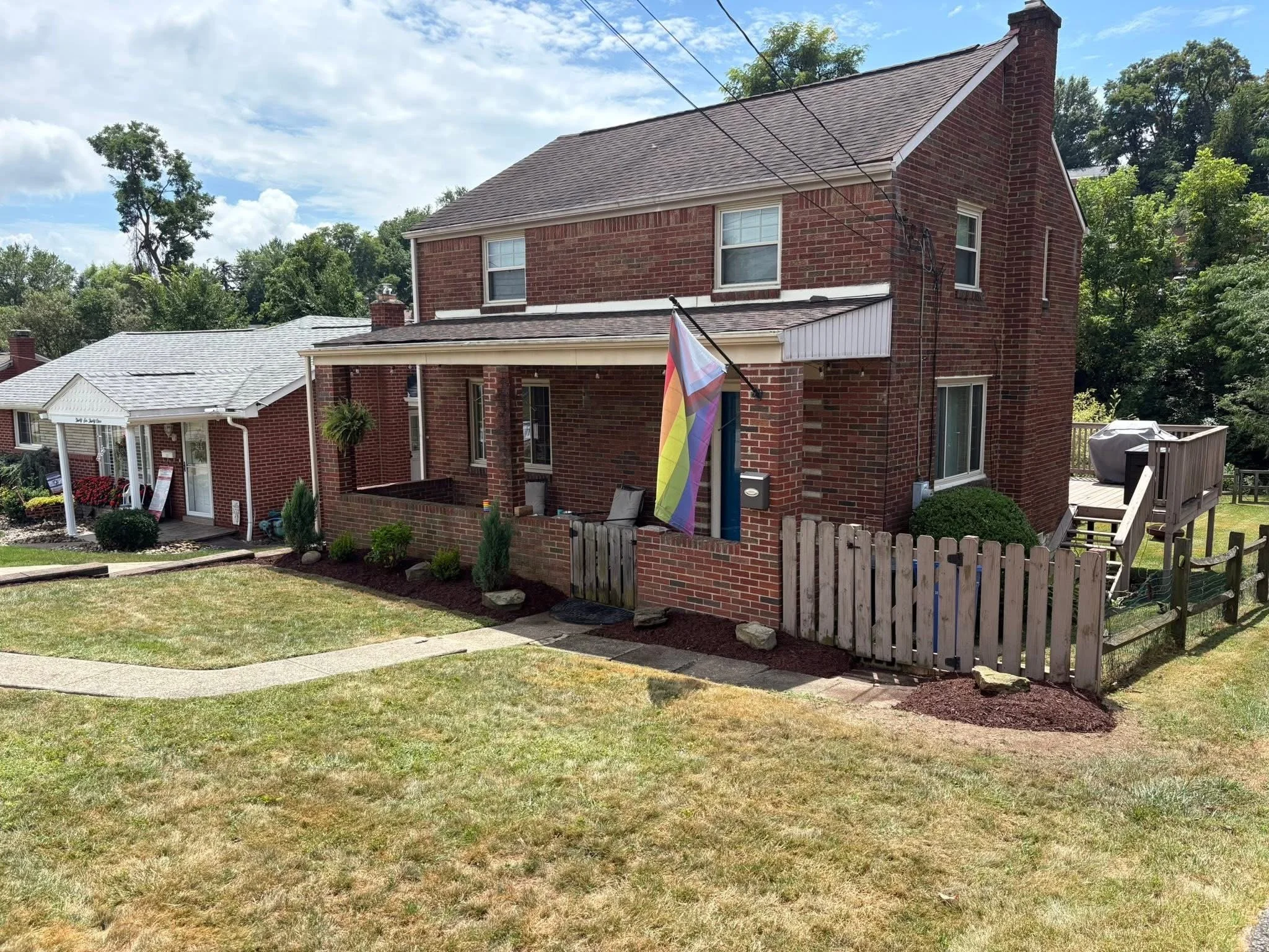Backyard of a red brick house with a porch, a rainbow flag, and a small fenced yard with stairs leading up to a deck. Neighboring house, trees, and a blue sky with clouds are also visible.