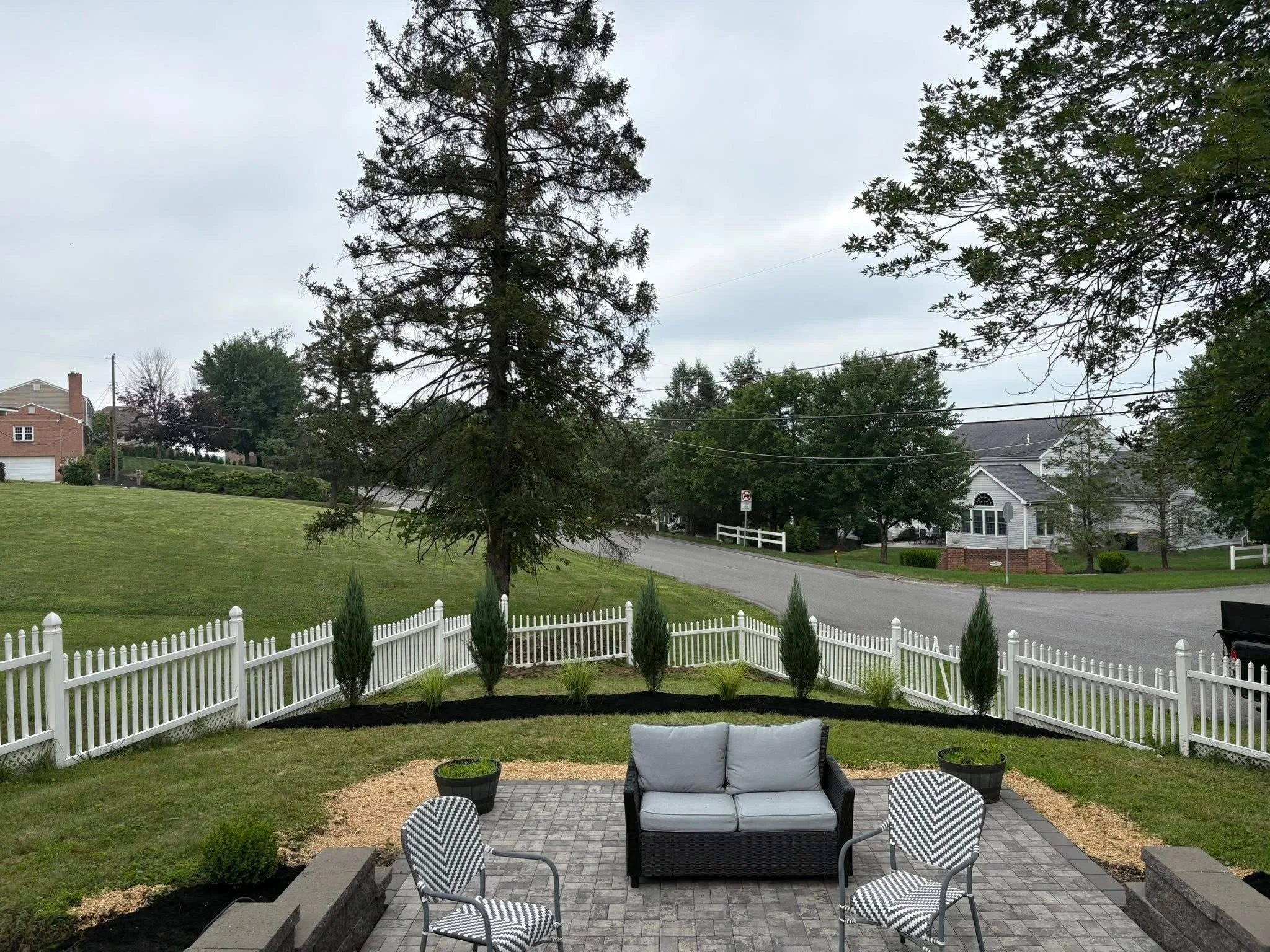 View of a backyard patio with a black wicker loveseat with gray cushions, two black and white striped chairs, and potted plants on a paved surface. A white picket fence surrounds the yard, with a large tree and green grass patches, and houses across 