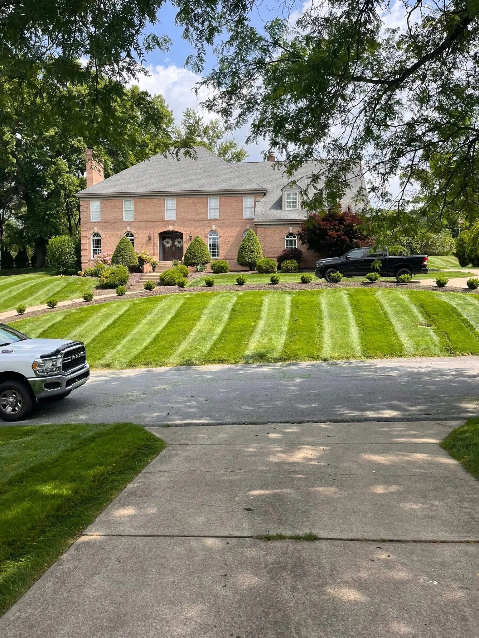 View of a large brick house with a well-manicured lawn and multiple vehicles parked in the driveway and on the street. The house is surrounded by trees and shrubs.