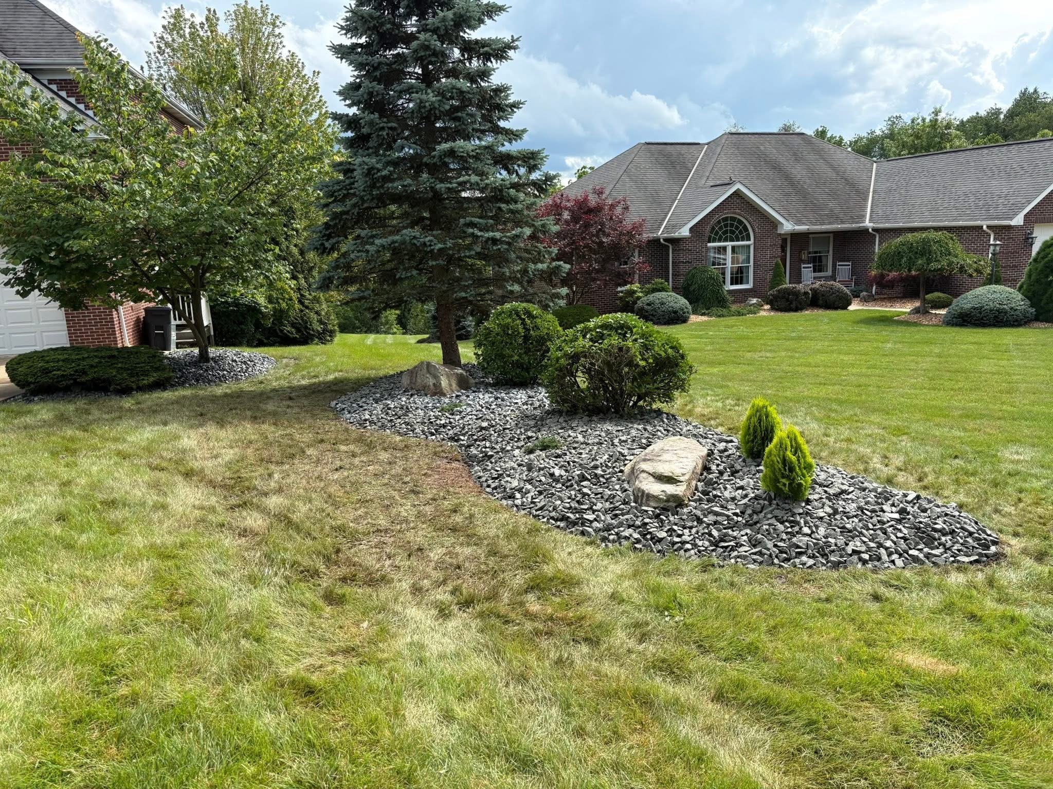 A well-maintained front yard with manicured green grass, decorative bushes, trees, and a landscaped rock garden in front of a brick house with a large window and a covered porch.