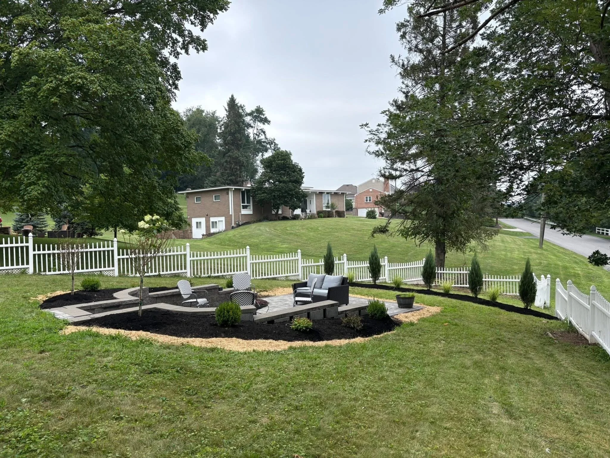 A backyard patio area with outdoor furniture, surrounded by a white picket fence and landscaped with small trees and shrubs on a grassy hill.