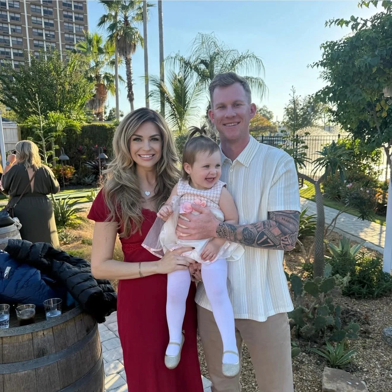 A smiling family of three standing in a garden, with a woman in a red dress, a man in a striped shirt, and a young girl in a dress and white tights, holding a happy baby girl.