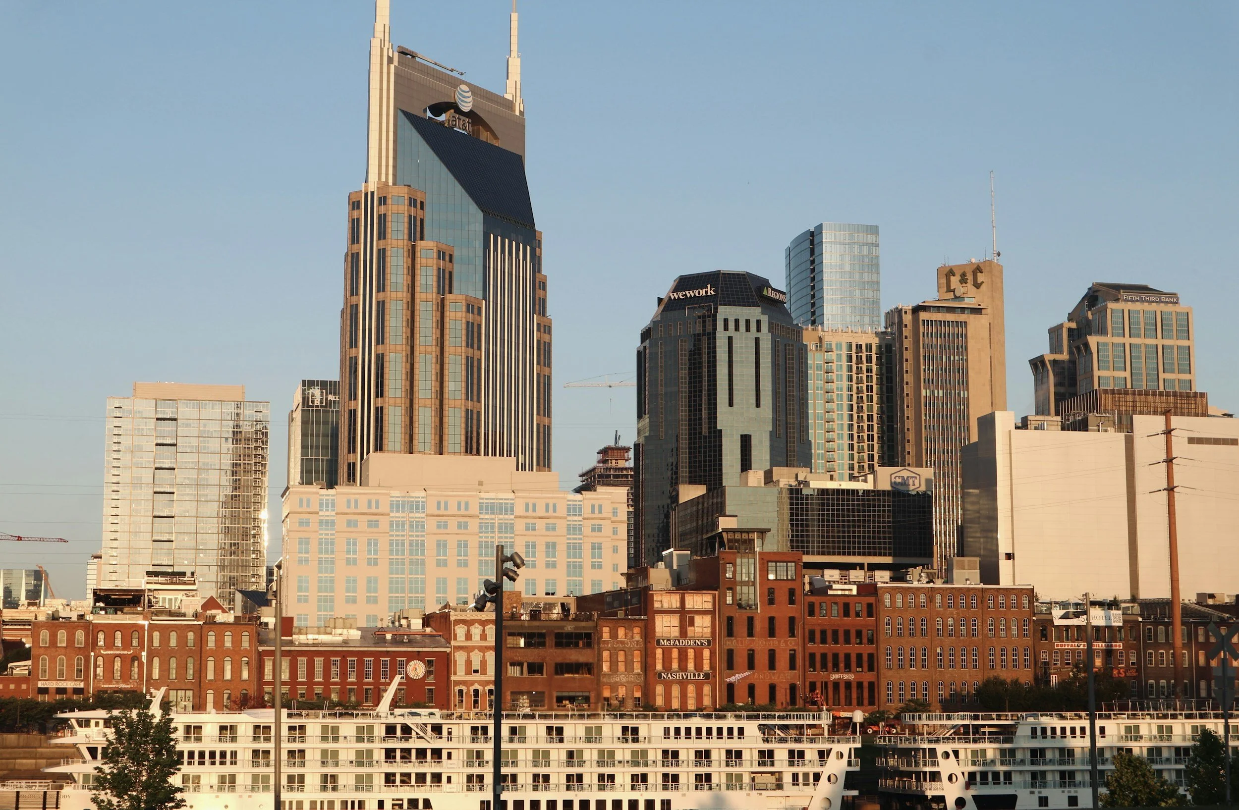 Downtown Nashville skyline with tall modern skyscrapers, historic brick buildings, and a river with a docked cruise ship in the foreground, taken during daylight hours.