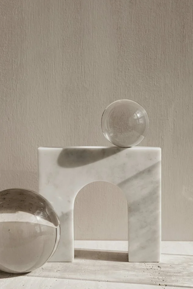 Two glass sphere sculptures, one on the ground and one on a marble arch, with shadows cast on a textured beige wall.