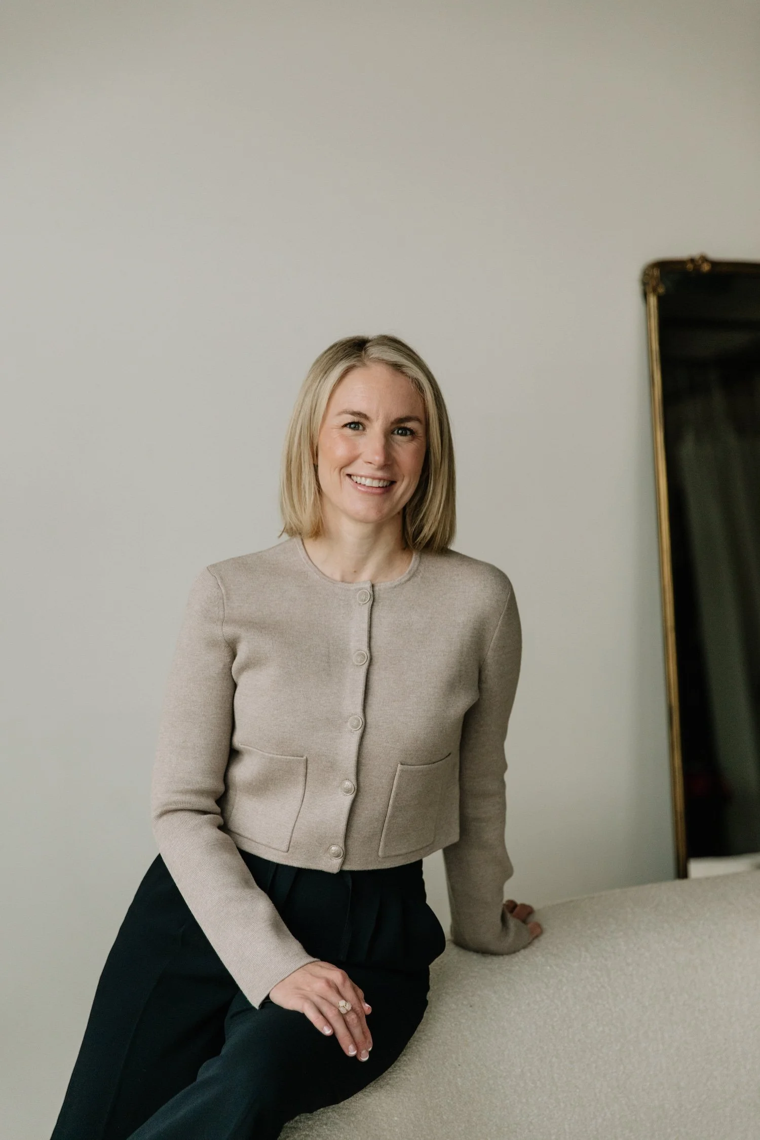 A woman dressed in a beige cardigan and black pants, smiling while leaning on a beige upholstered surface, with a large mirror with a gold frame in the background.