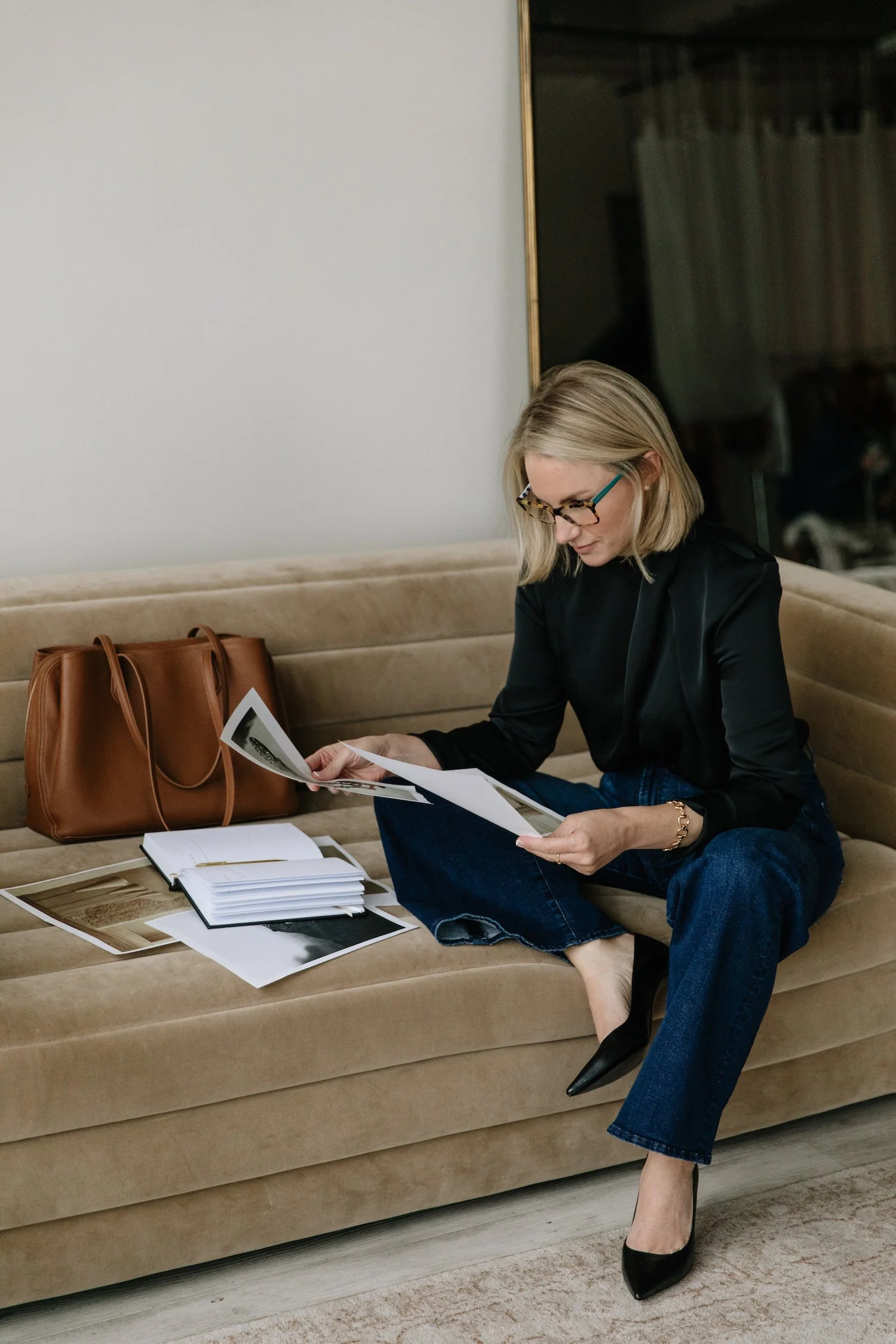 A woman with blonde hair, black top, blue jeans, and black high heels sitting on a beige sofa, reviewing photographs and documents with a brown purse nearby and some open books and papers on the sofa.