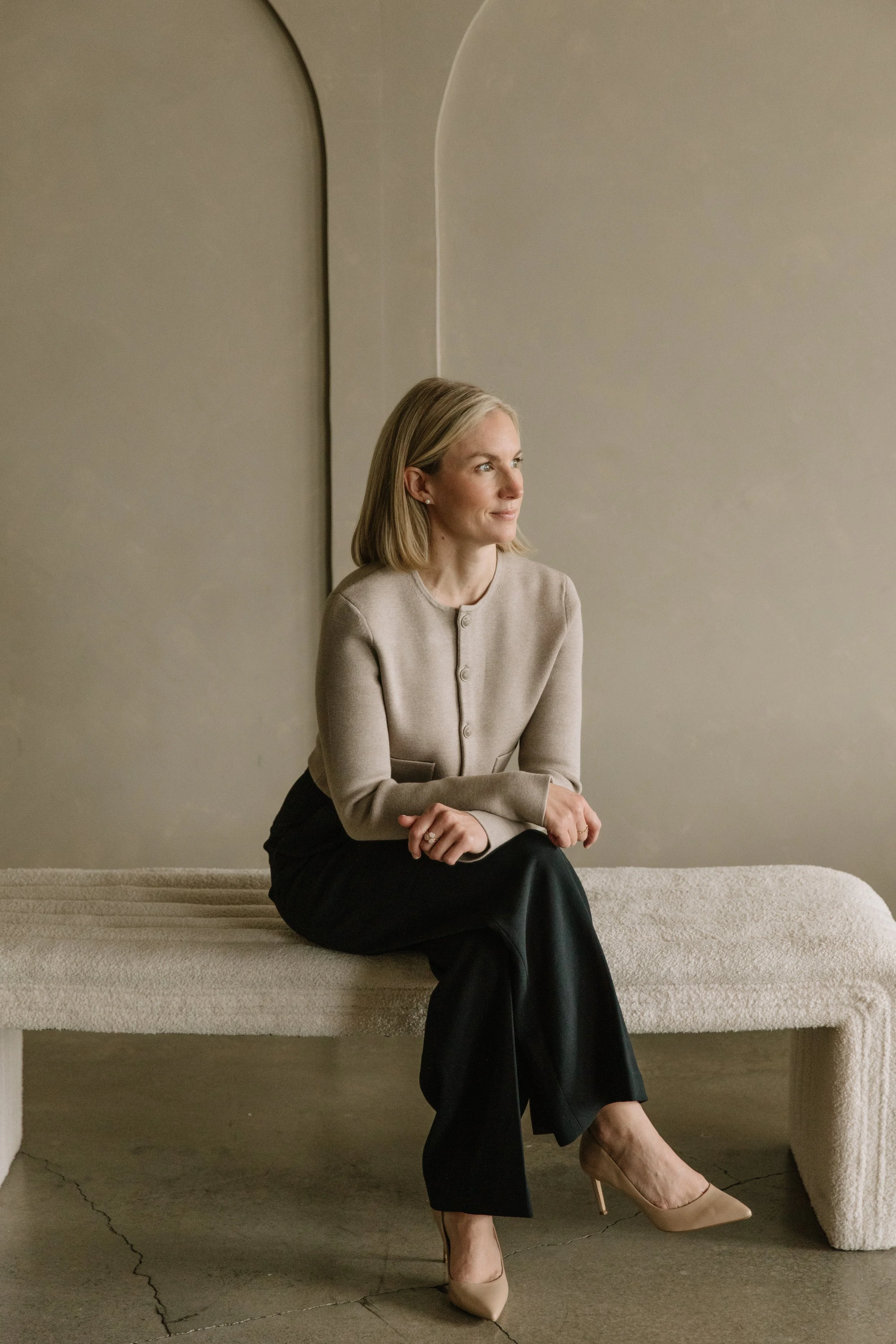 A woman with blonde hair sitting on a beige textured bench, looking to her right with a calm expression. She is dressed in a beige buttoned top, black pants, and beige high heels, and is set against a neutral beige wall background.