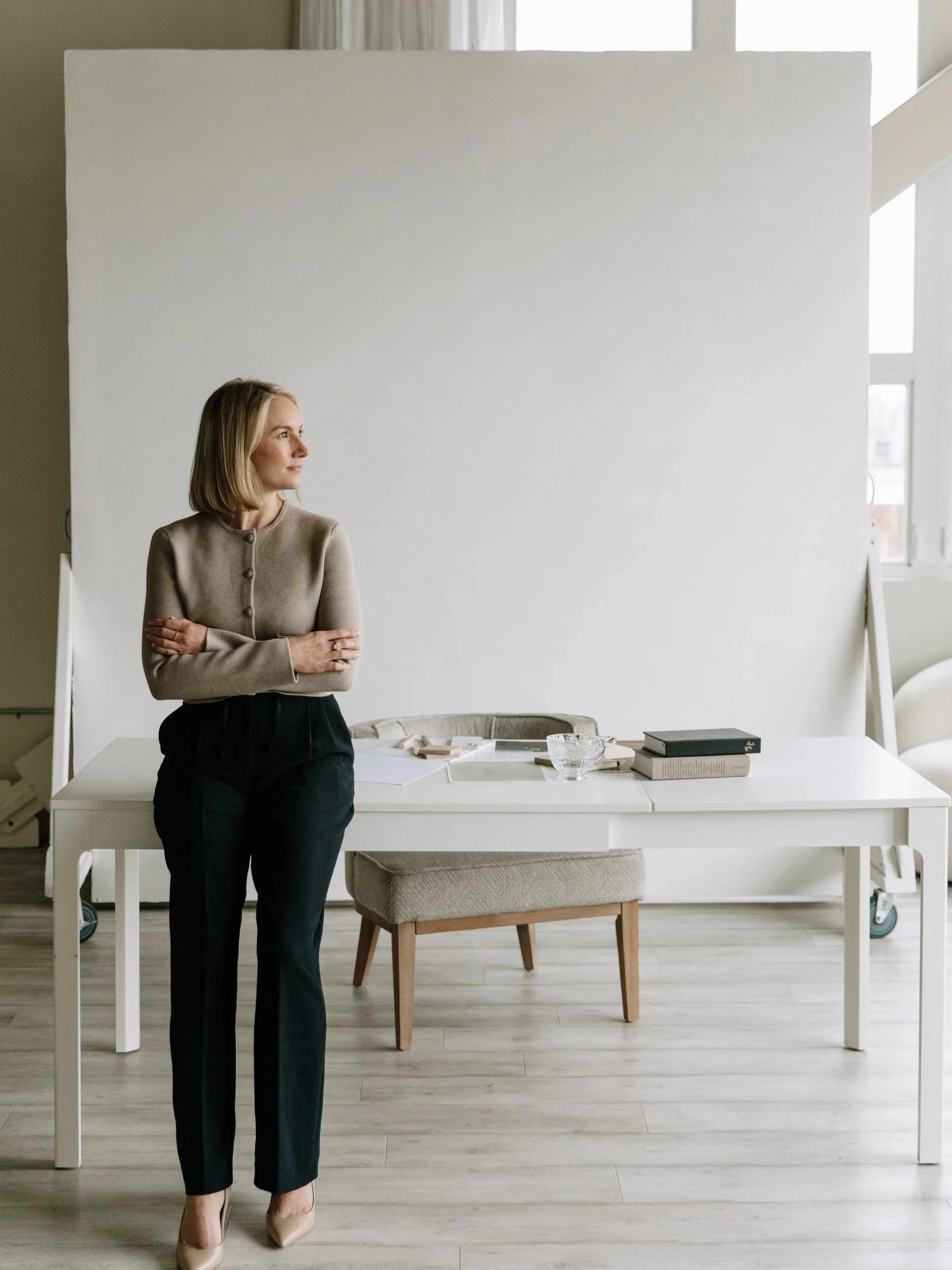 A woman with shoulder-length blonde hair, wearing a tan cardigan and black trousers, stands with crossed arms in a bright room with natural light. Behind her is a white table with books and glassware, and a chair with beige upholstery. The background includes a large blank whiteboard or backdrop and windows.