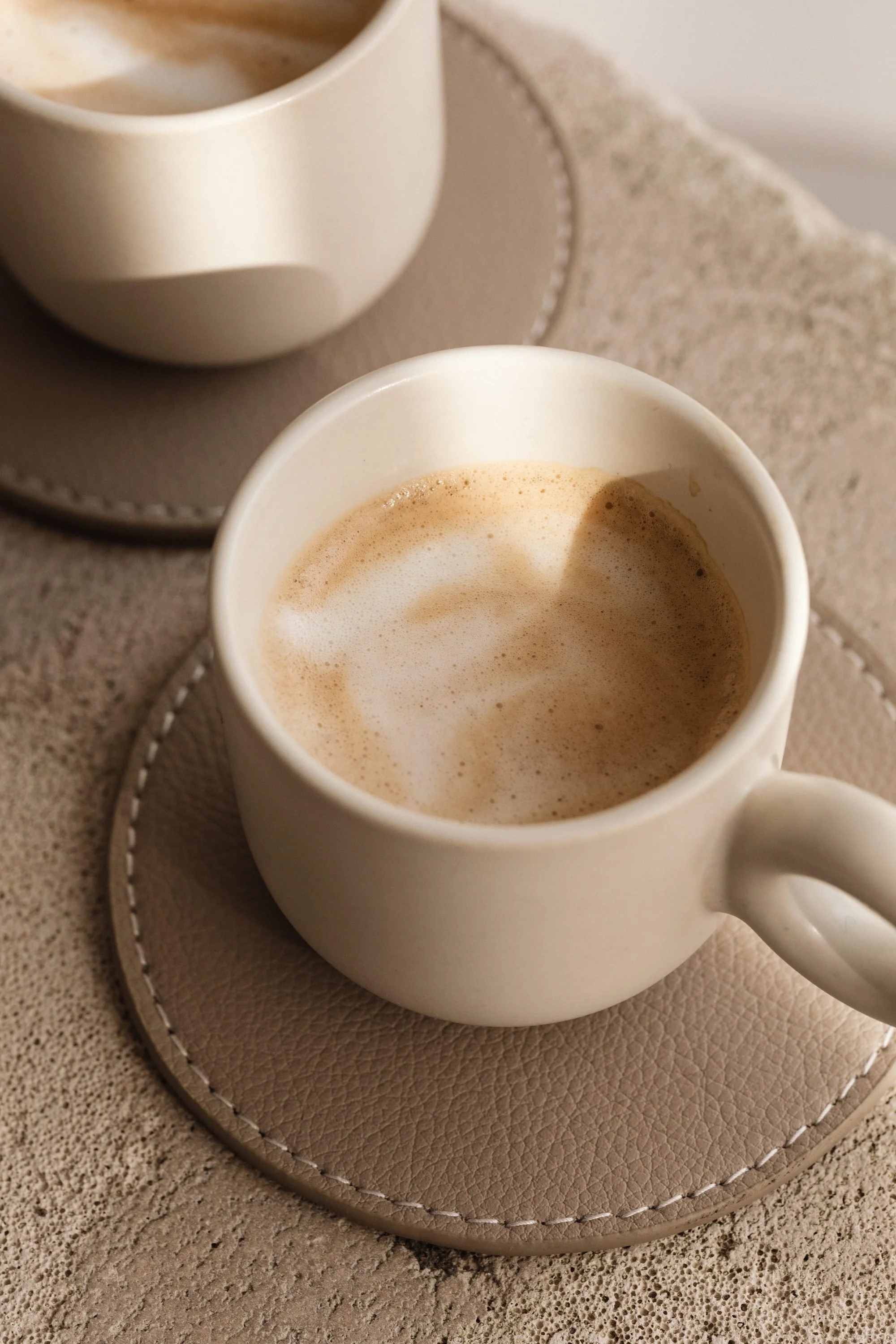 A white ceramic mug filled with coffee topped with frothy milk, placed on a beige leather coaster on a textured beige surface.