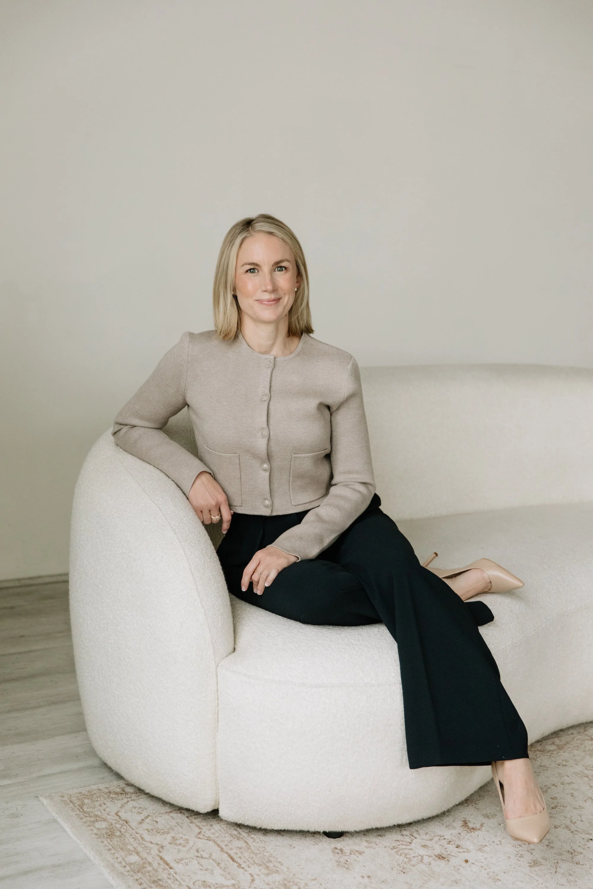 A woman with blonde hair wearing a beige cardigan, black pants, and nude high heels sitting on a white curved sofa in a room with light-colored walls and wooden flooring.