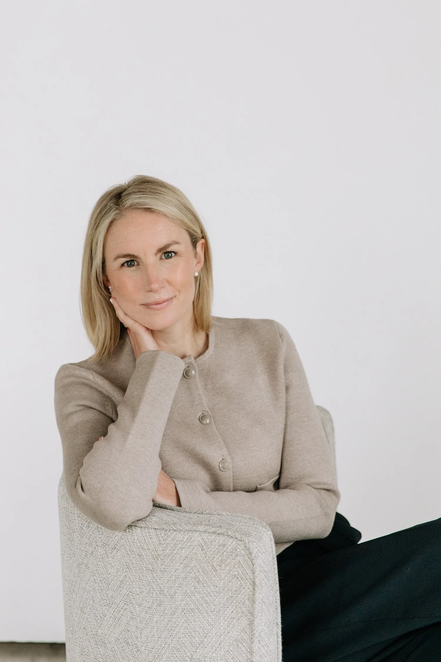 A woman with blonde hair sitting on an off-white textured armchair, wearing a beige button-up top and black pants, resting her face on her hand and smiling softly at the camera against a plain white background.