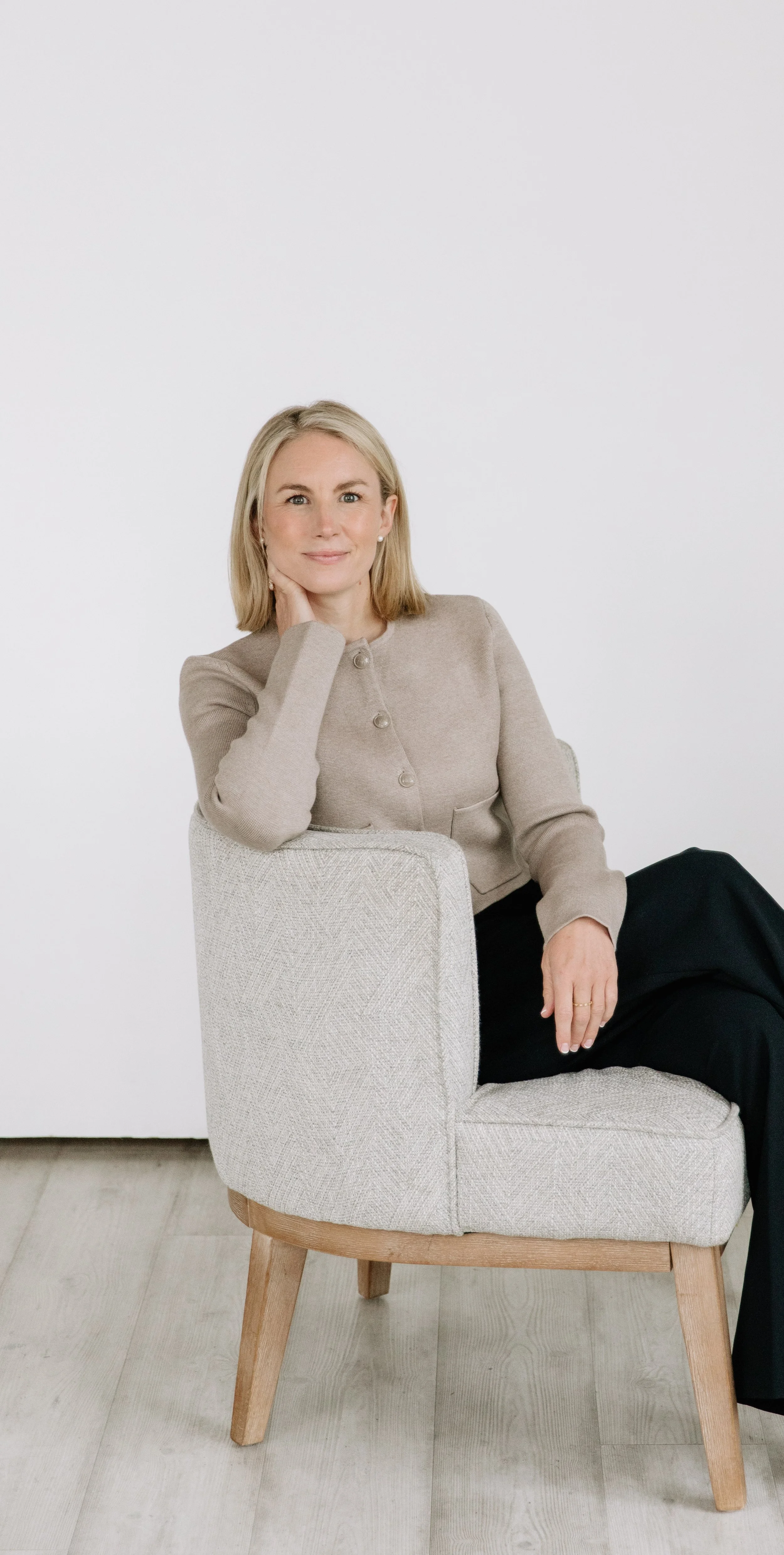 A woman with blonde hair, wearing a beige blazer, seated on a light-colored armchair with wooden legs, against a plain white background.