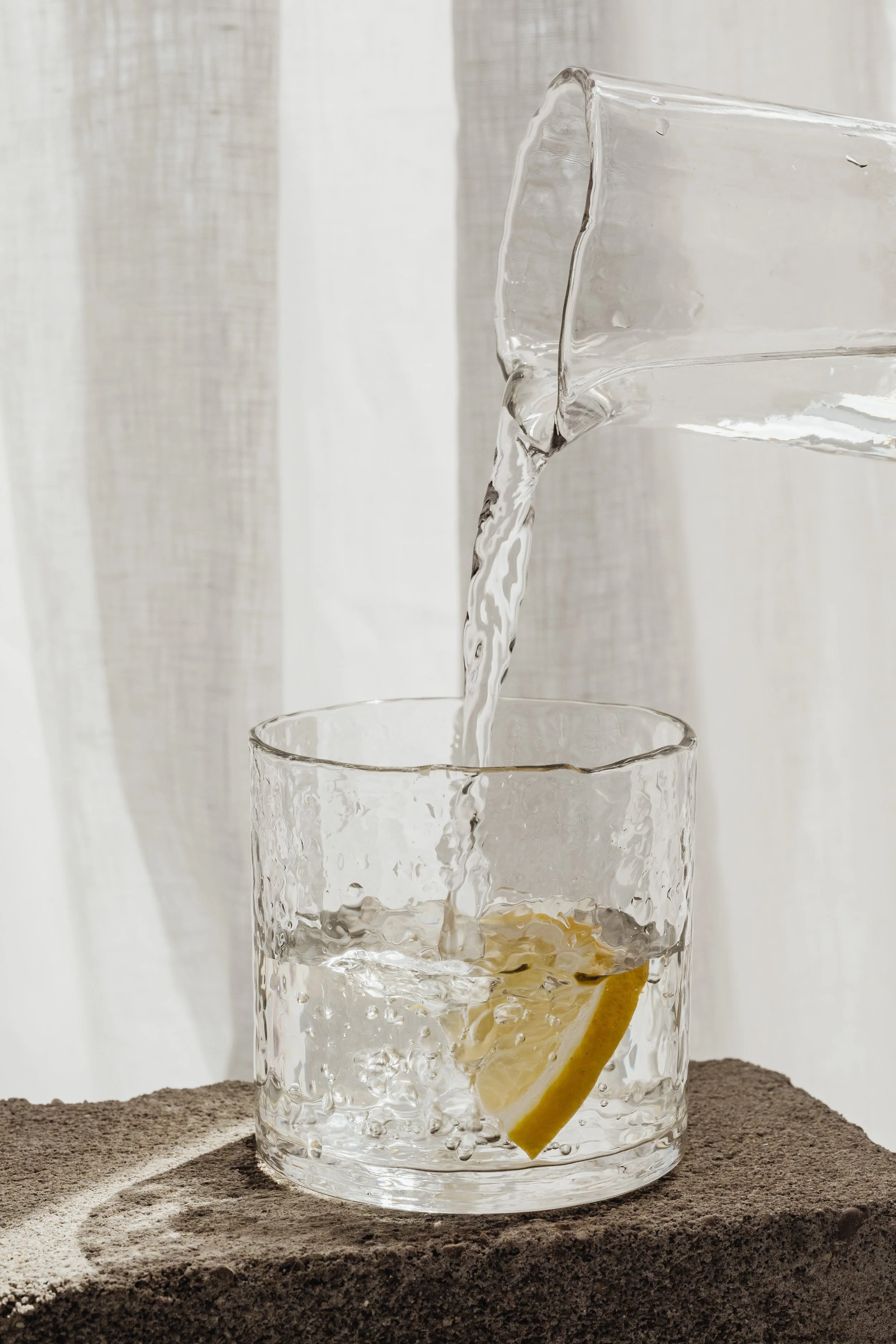 A clear glass with a lemon wedge inside, placed on a textured surface, with water being poured into it from a glass pitcher.