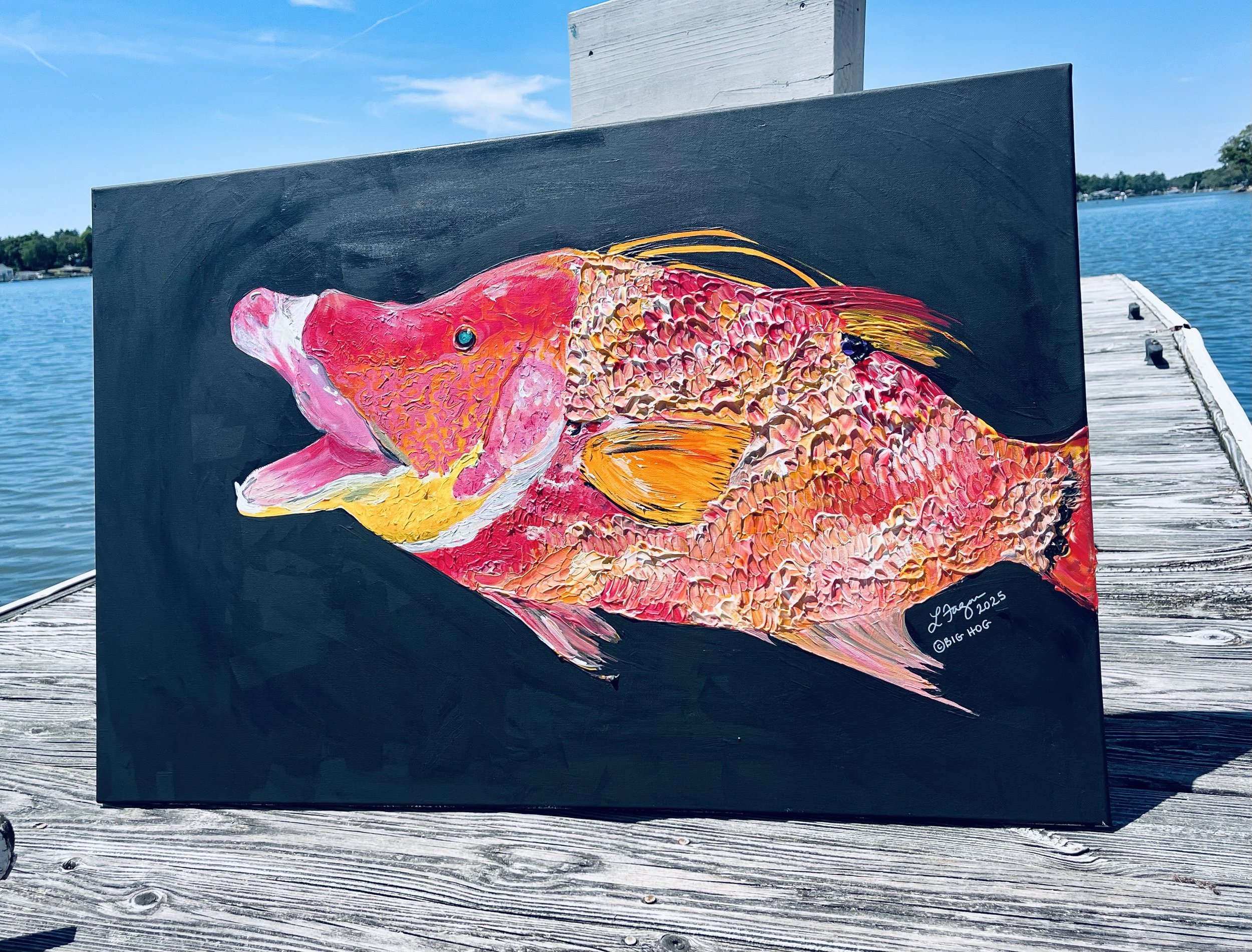 Painting of a pink and orange fish on a black background, displayed on a wooden dock near a body of water.