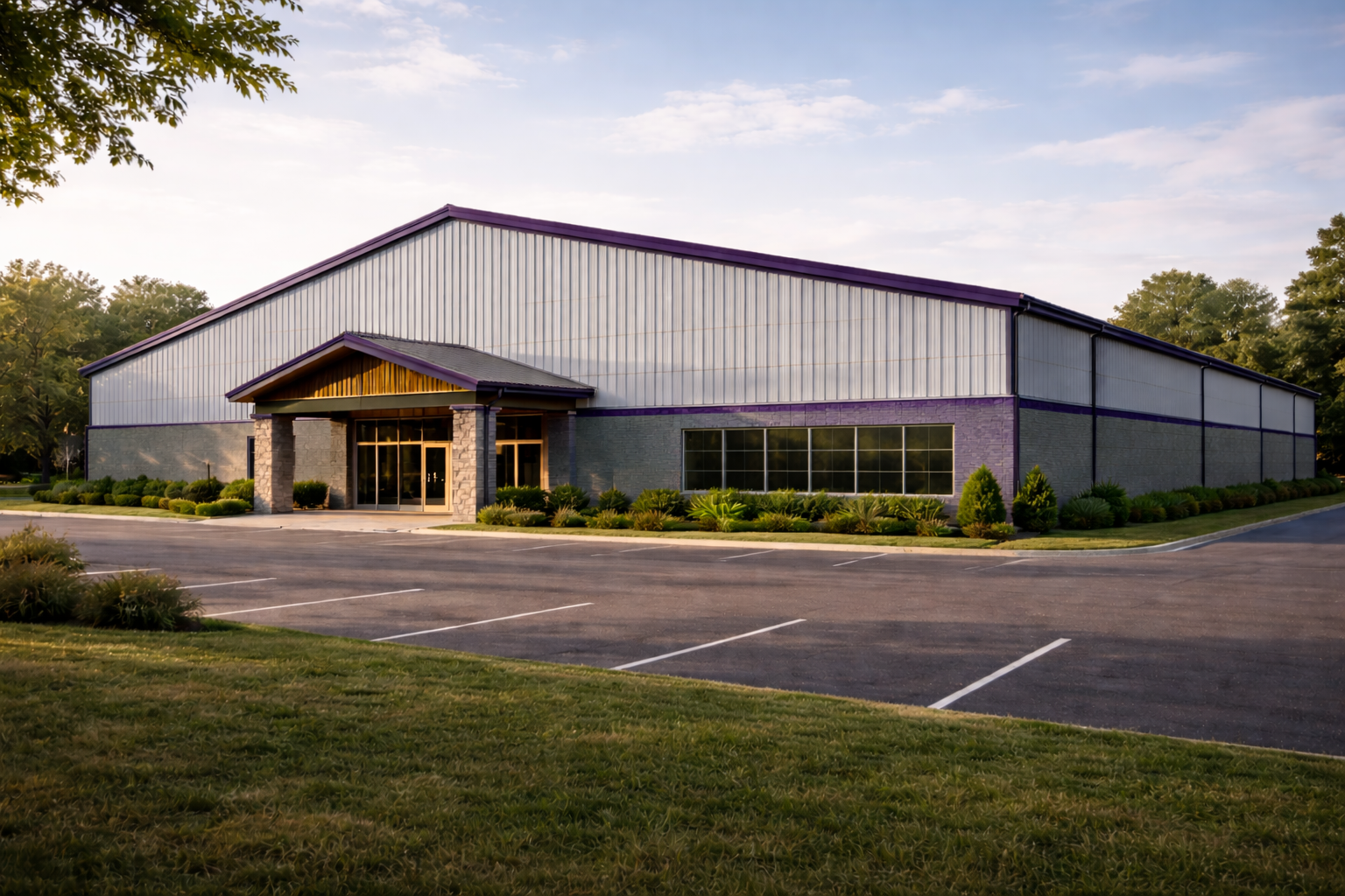 A Indoor Tennis building with metal siding, purple trim, and a peaked roof, surrounded by a landscaped area with bushes and trees, and an empty parking lot.