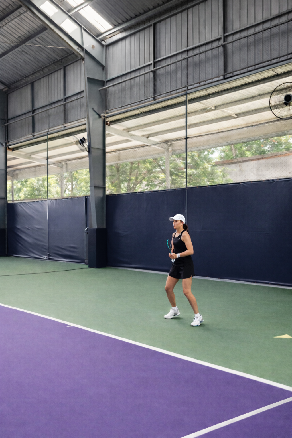A woman in black tennis attire and white cap standing on a tennis court holding a tennis racket inside an indoor tennis facility.