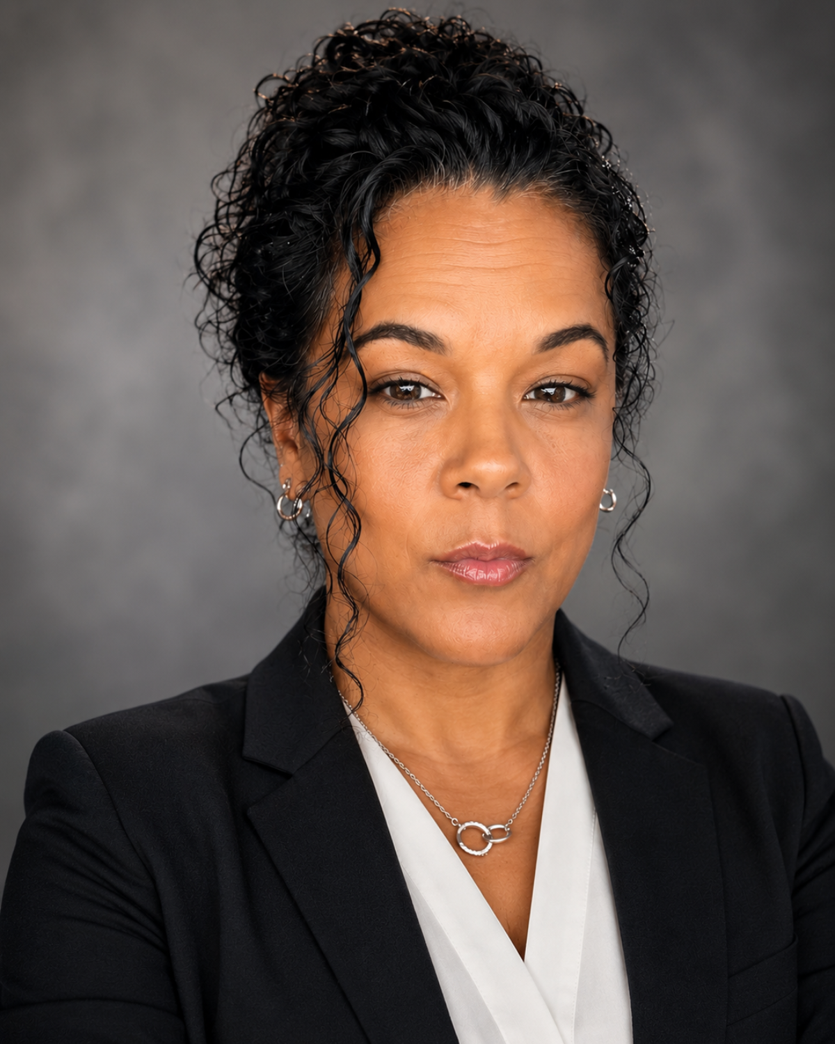 A woman with medium skin tone, curly black hair, and wearing a black blazer and white blouse, looking confident against a gray background.