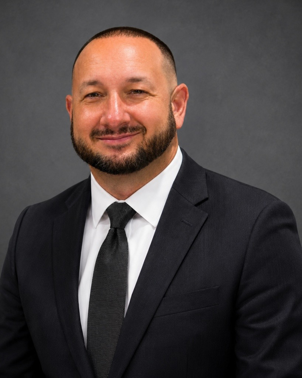 A man in a black suit, white shirt, and black tie, smiling, with a beard and short hair, against a gray background.