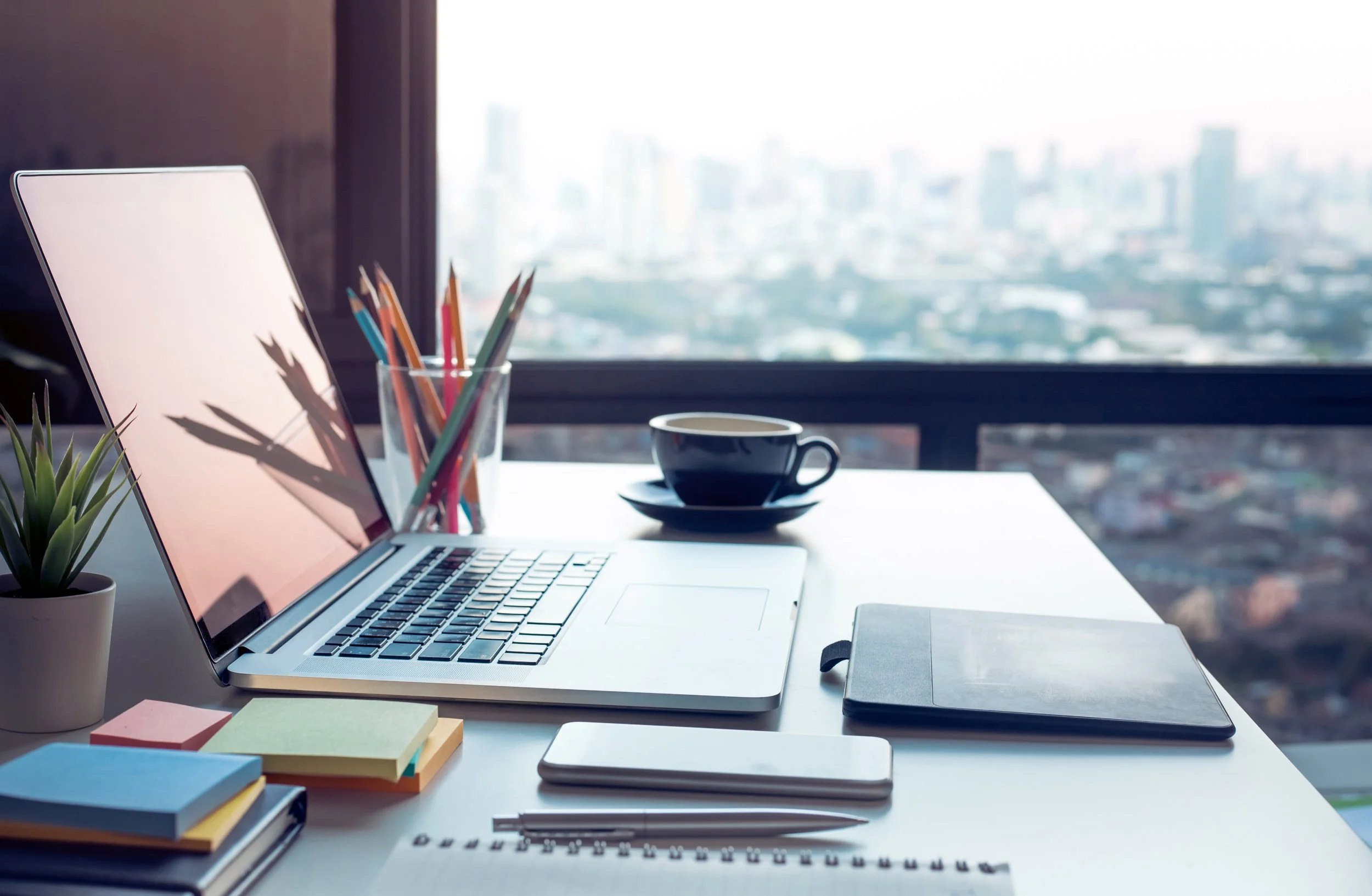 A modern workspace with a silver laptop, a potted succulent, colorful pencils in a glass, a white cup with dark liquid, notebooks, sticky notes, a mouse, and a large window showing a cityscape in the background.