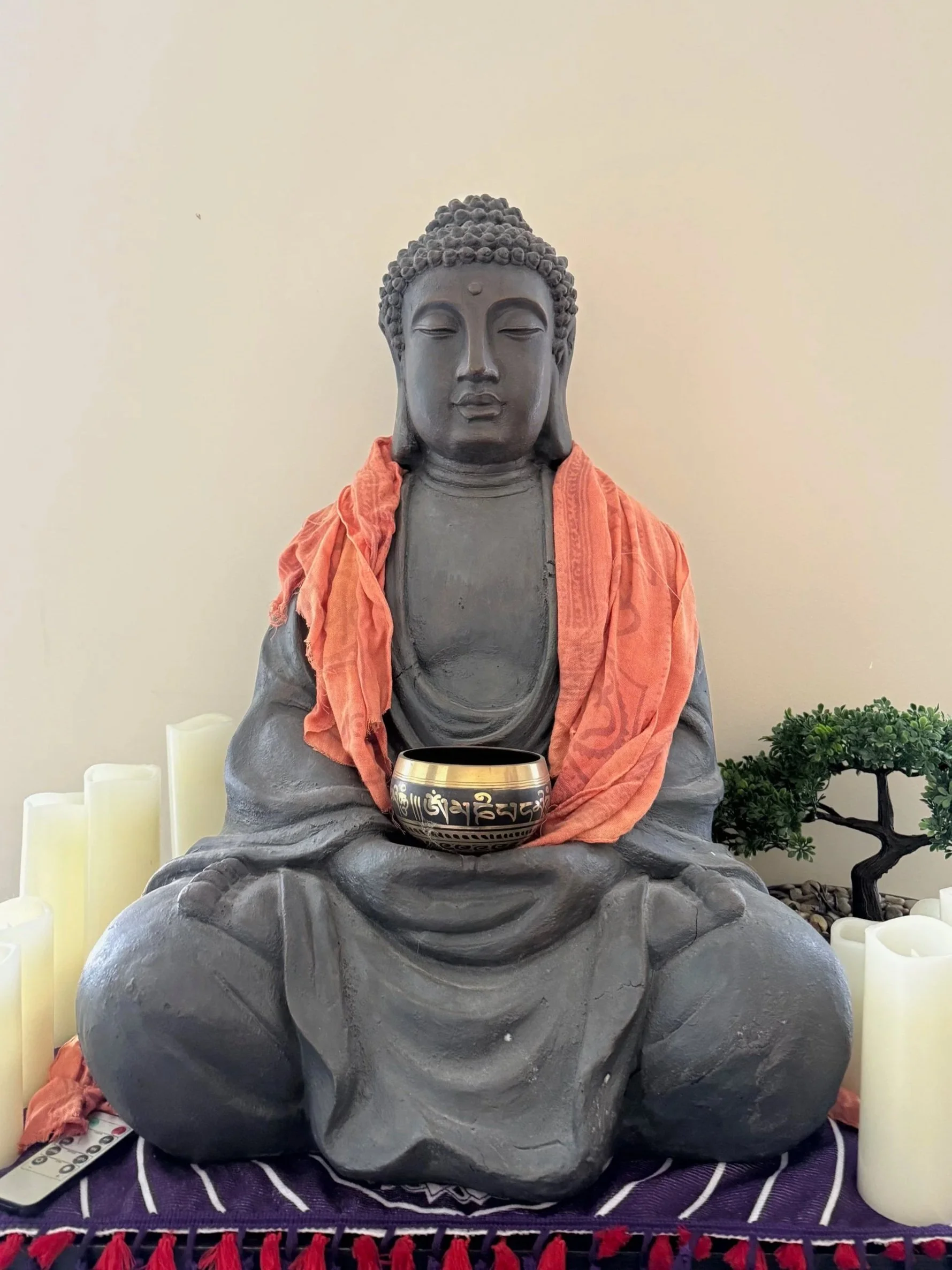 A black stone Buddha statue in a seated meditation pose, adorned with an orange cloth around the neck. The statue holds a bowl with inscriptions in an unknown language in its lap. There are white candles on either side, a small tree on the right, and a purple cloth with red tassels underneath.