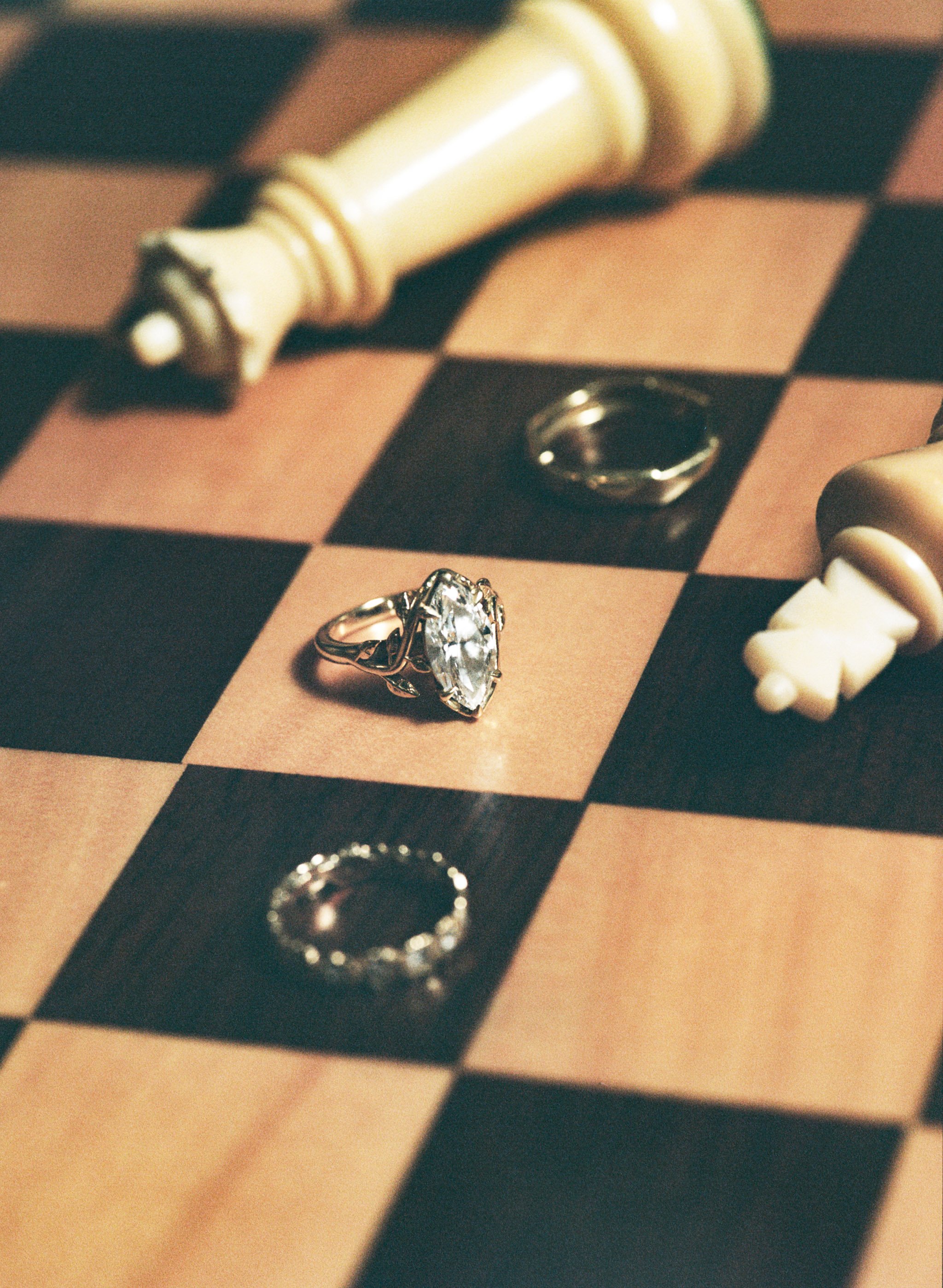Close-up of a chessboard with a diamond ring, sitting on the black and brown checkered surface, surrounded by chess pieces, including a king, queen, and a pawn.