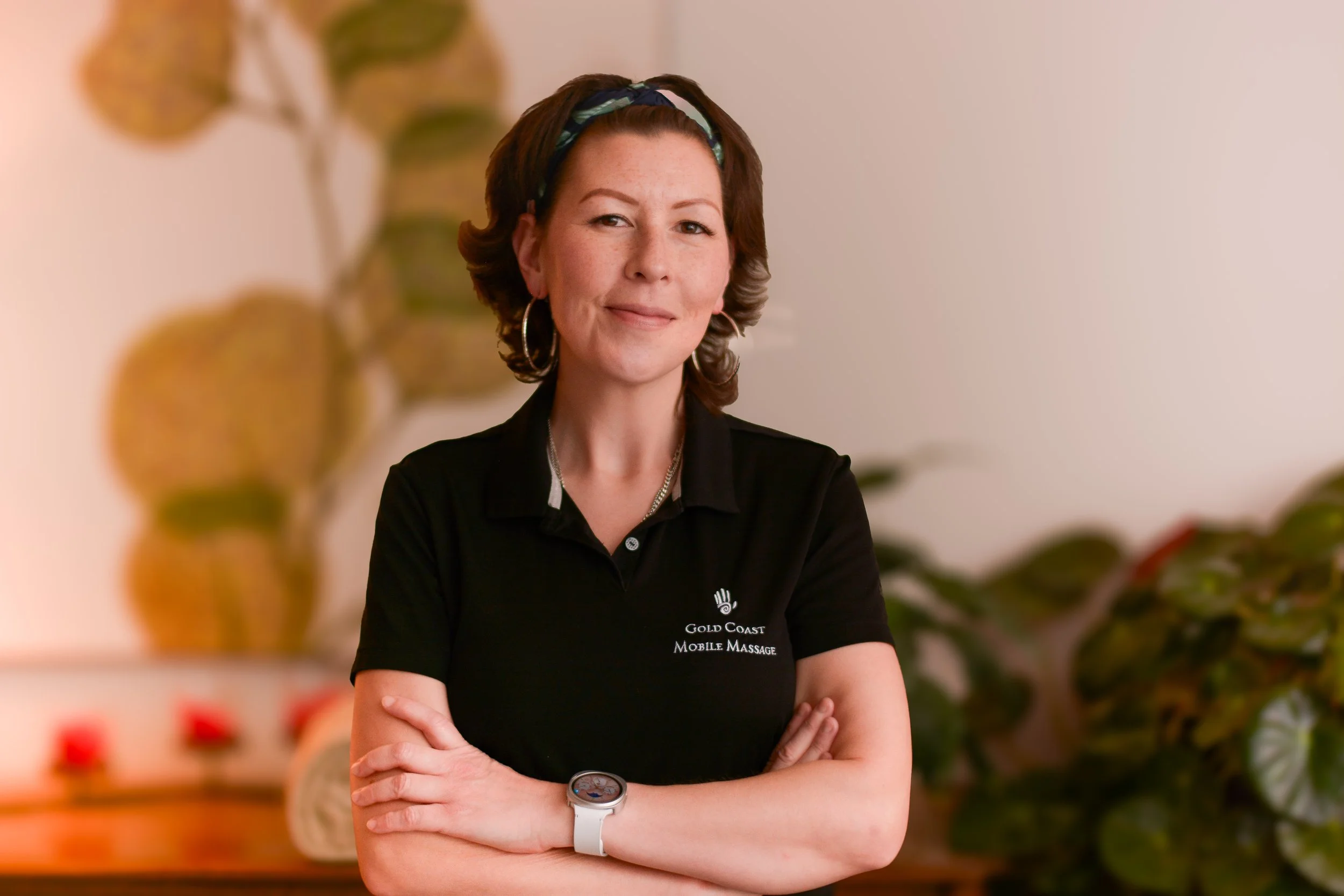 A woman with short brown hair, wearing a black polo shirt with a logo that says 'Gold Coast Mobile Massage,' standing with her arms crossed and smiling, indoors with plants and soft lighting.