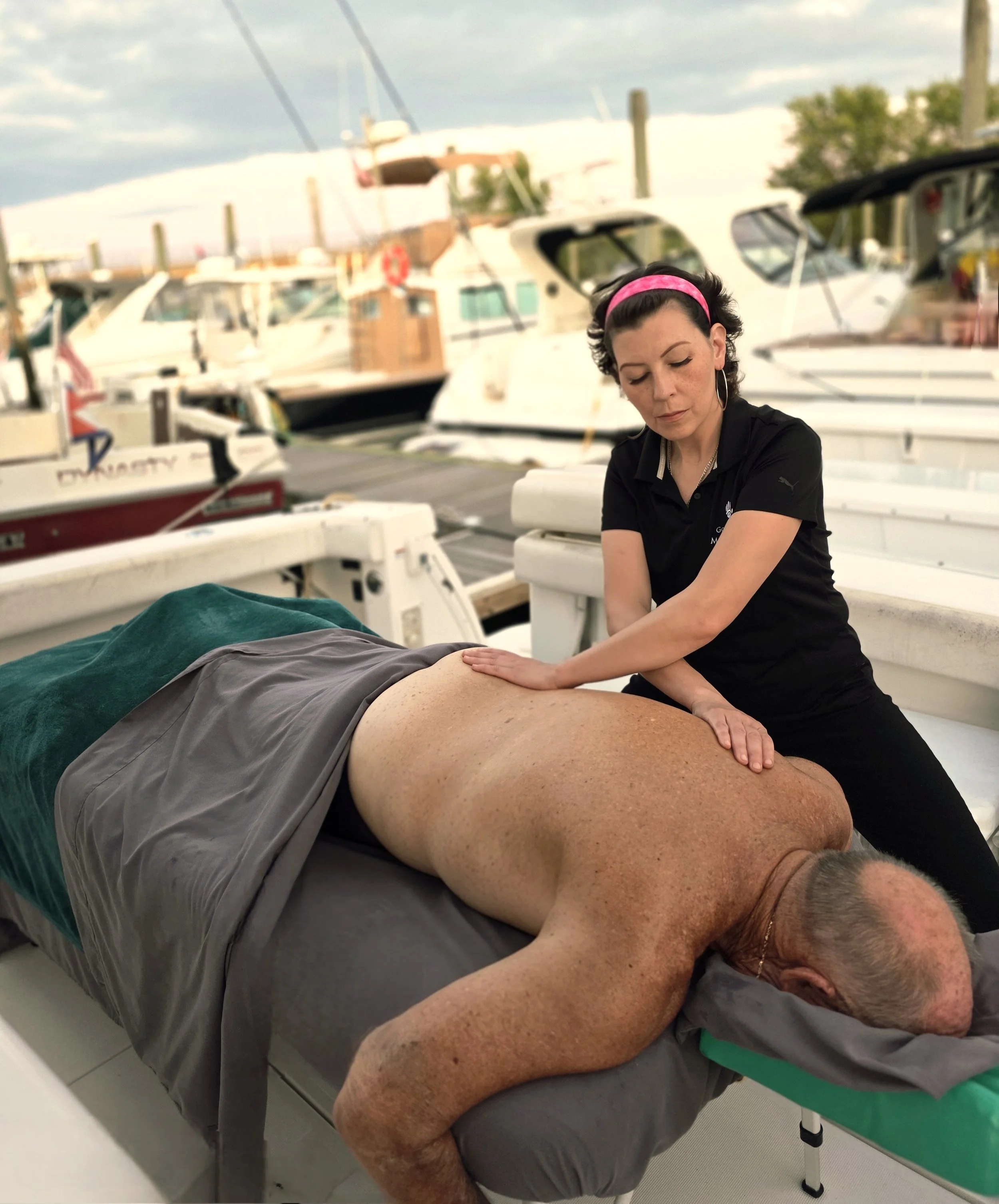 A woman giving a massage to a man lying face down on a table on a dock, with boats in the background.