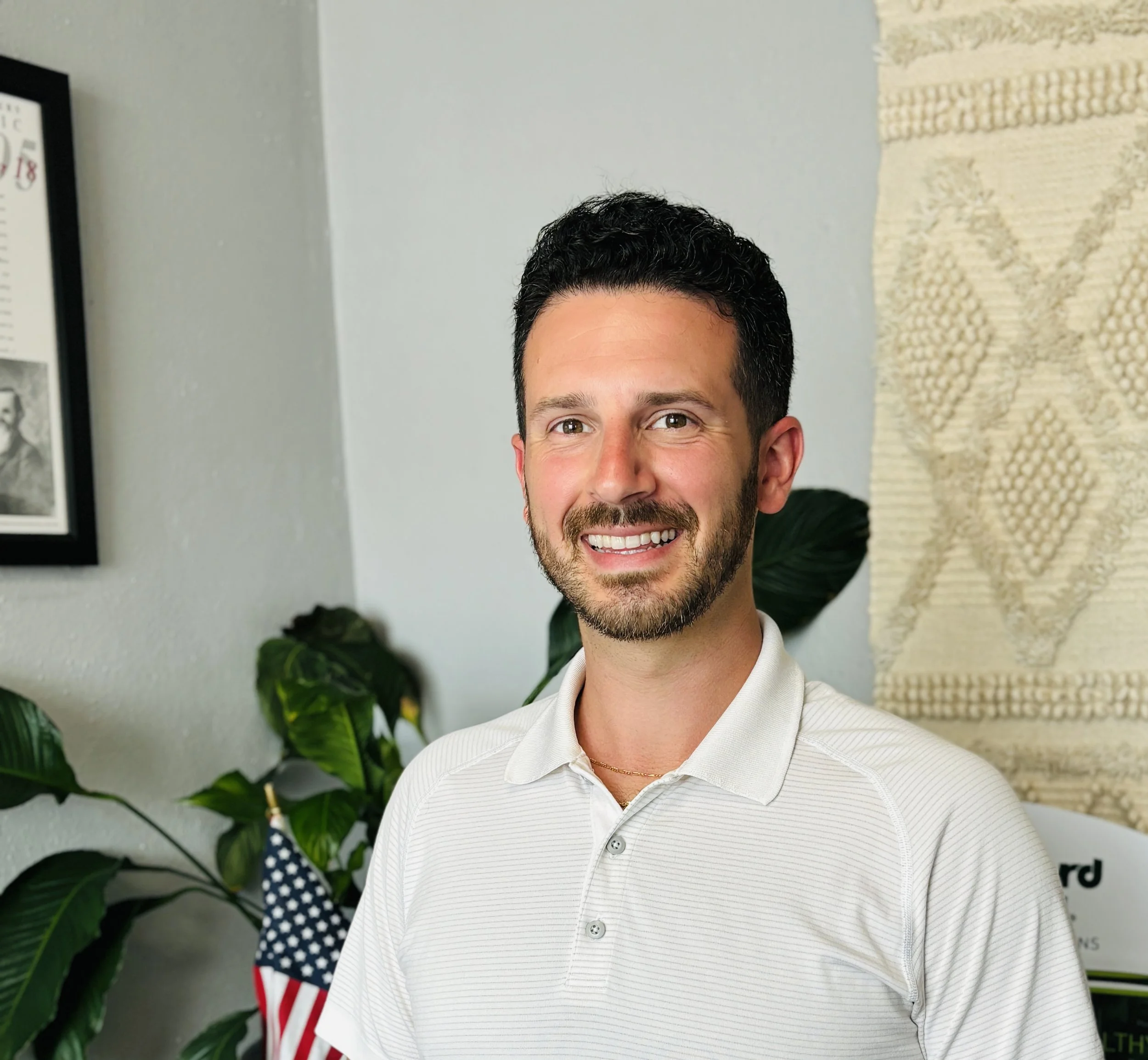 A smiling man with dark hair, beard, wearing a white collared shirt, standing indoors with plants and framed pictures and a wall hanging in the background.