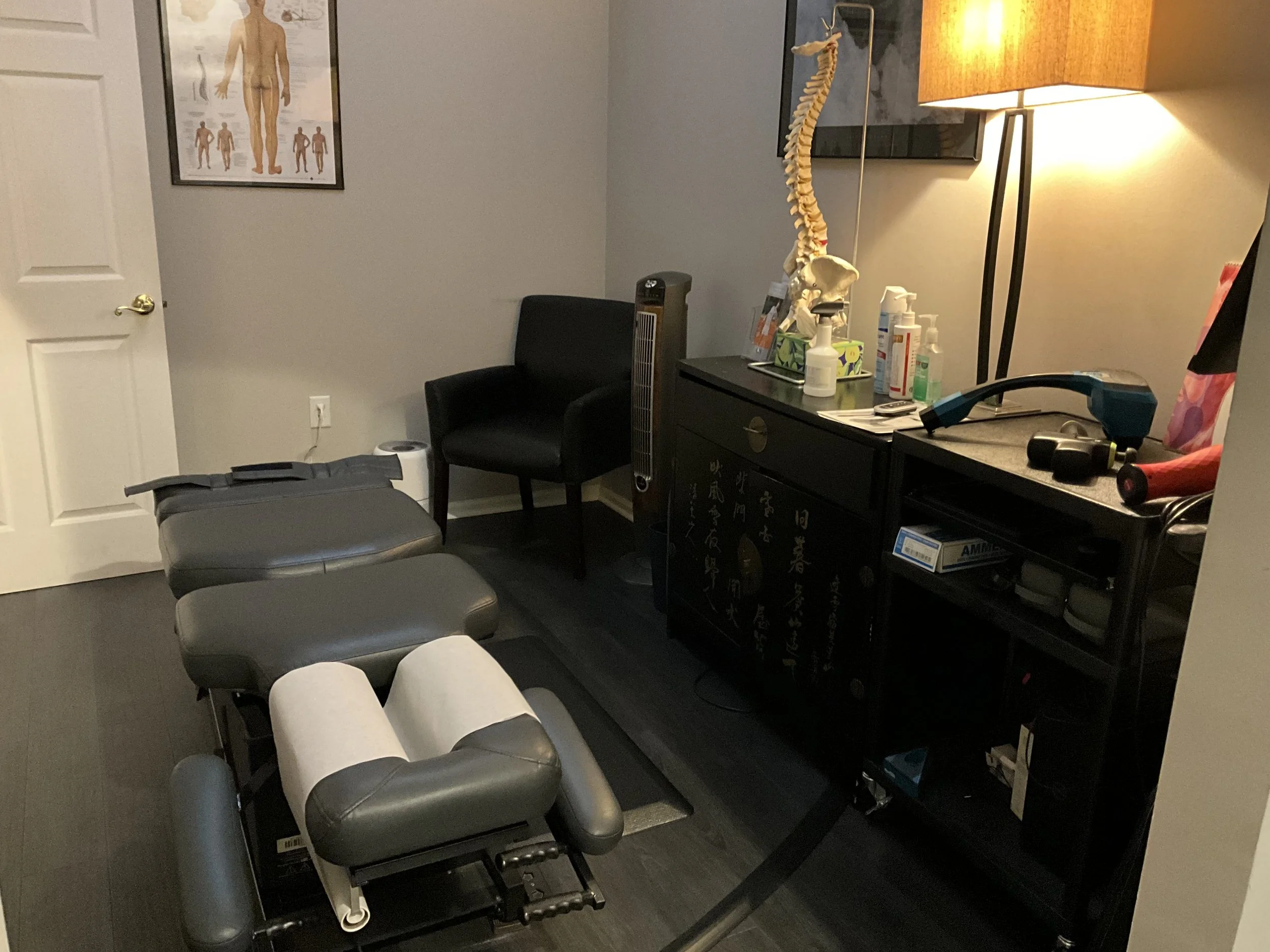 A chiropractic office room with a treatment table, a black chair, a cabinet with supplies, a lamp, a model of a human spine, and a poster of the human body on the wall.
