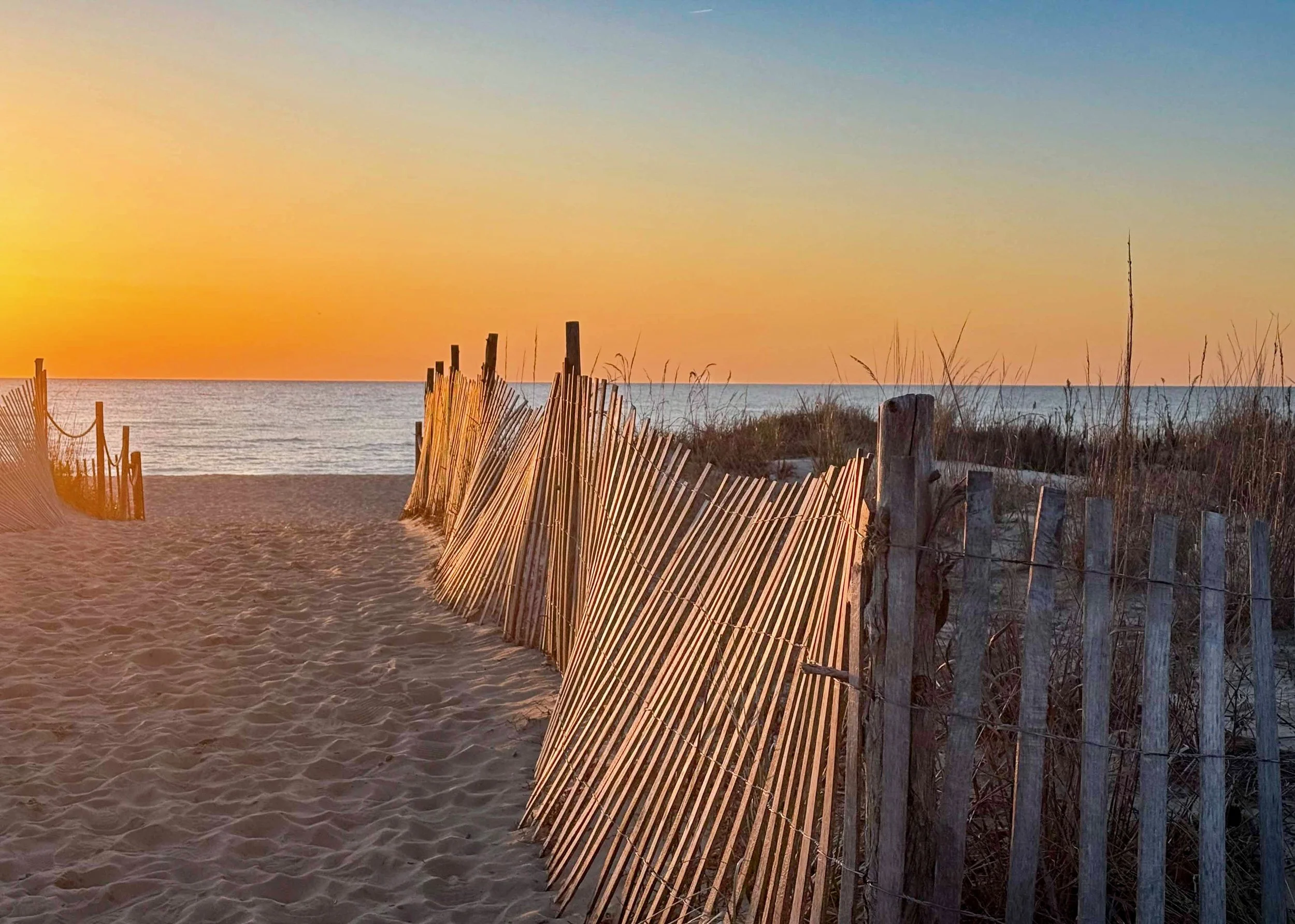 A sandy beach with a pathway leading to the ocean, lined with wooden and bamboo fences, during sunset with a clear sky.