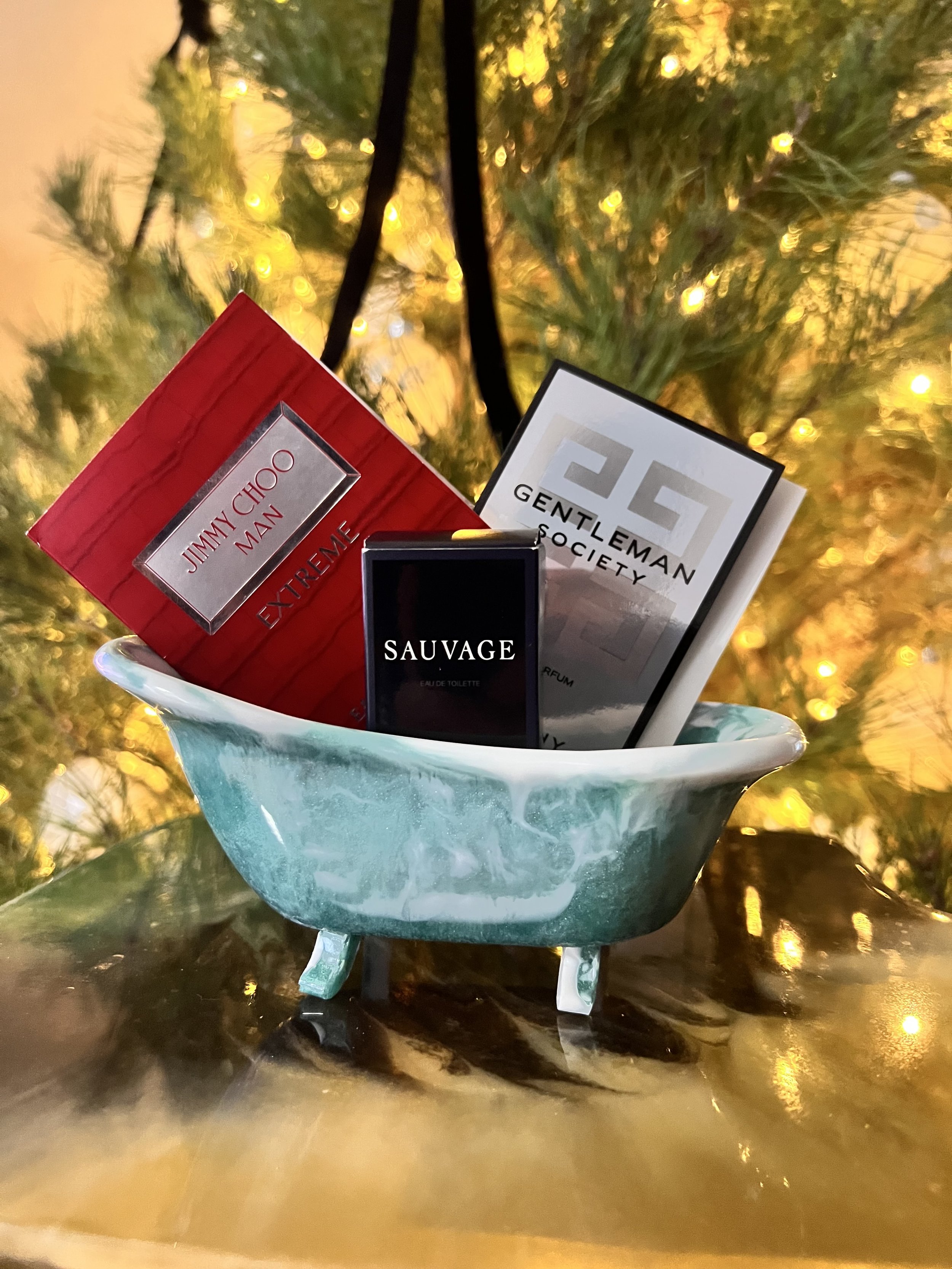 Miniature bathtub filled with perfume bottles and boxes in front of a Christmas tree with lights.