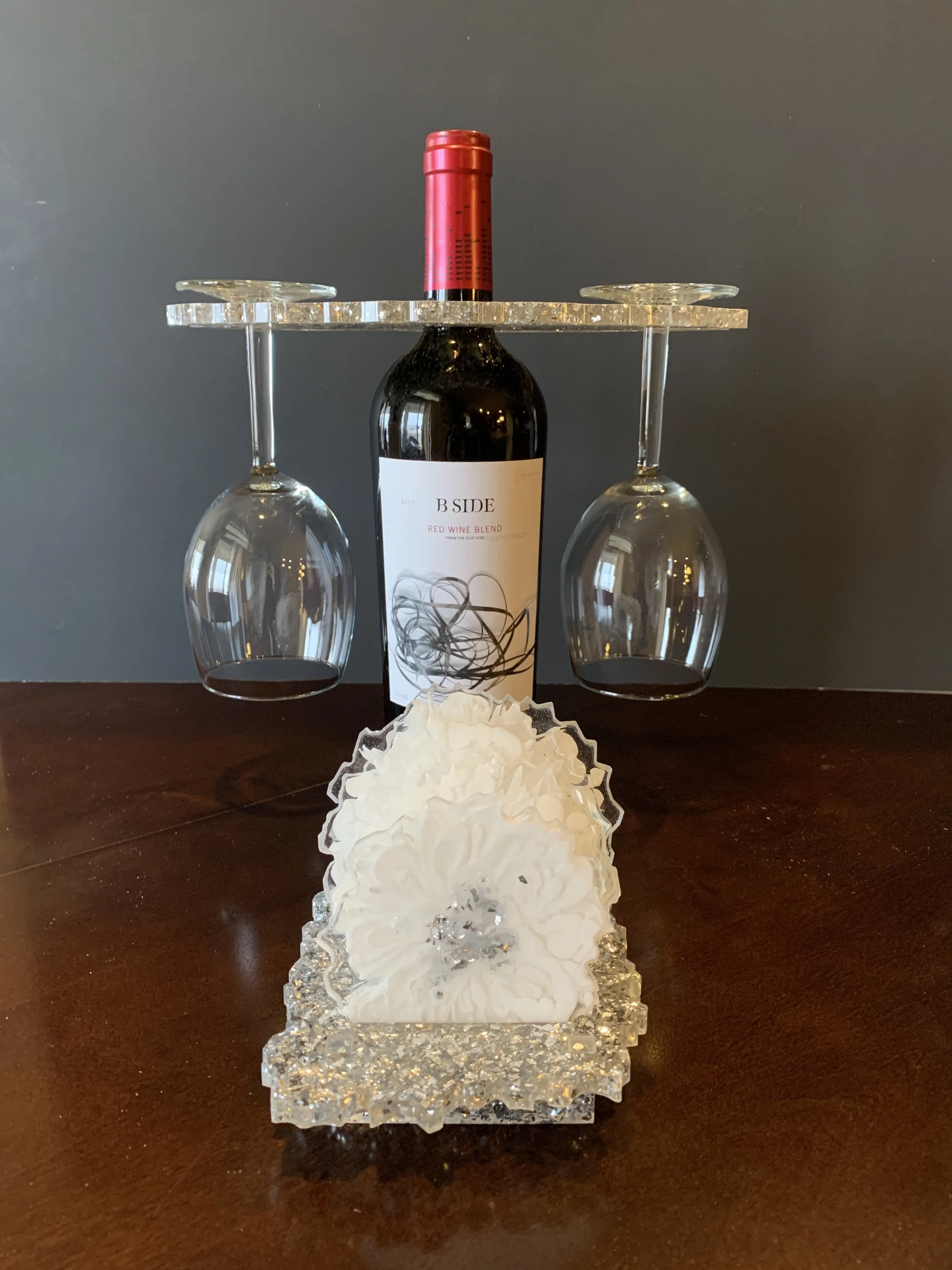 Decorative wine display with a wine bottle, two upside-down wine glasses, and a white decorative flower on a glass base in front of a dark background.