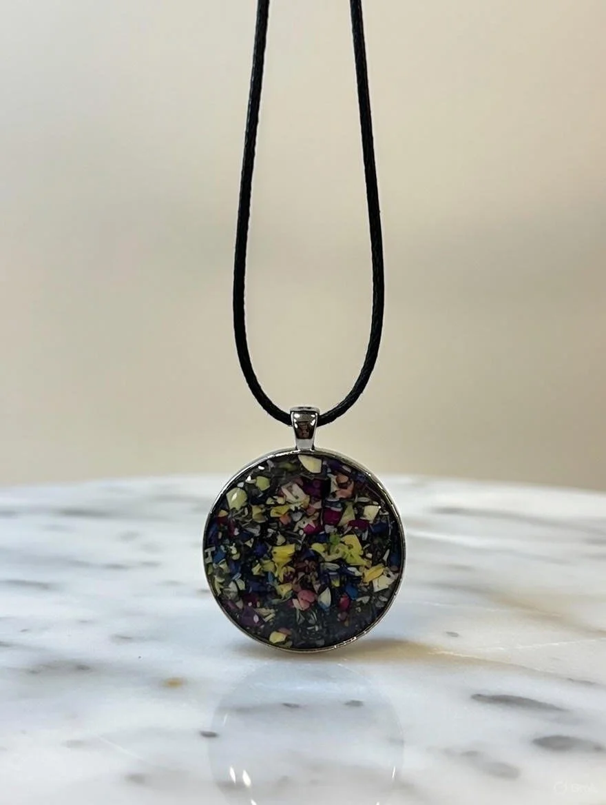 Round pendant necklace with colorful speckled mosaic design, hanging on a black cord against a neutral background.