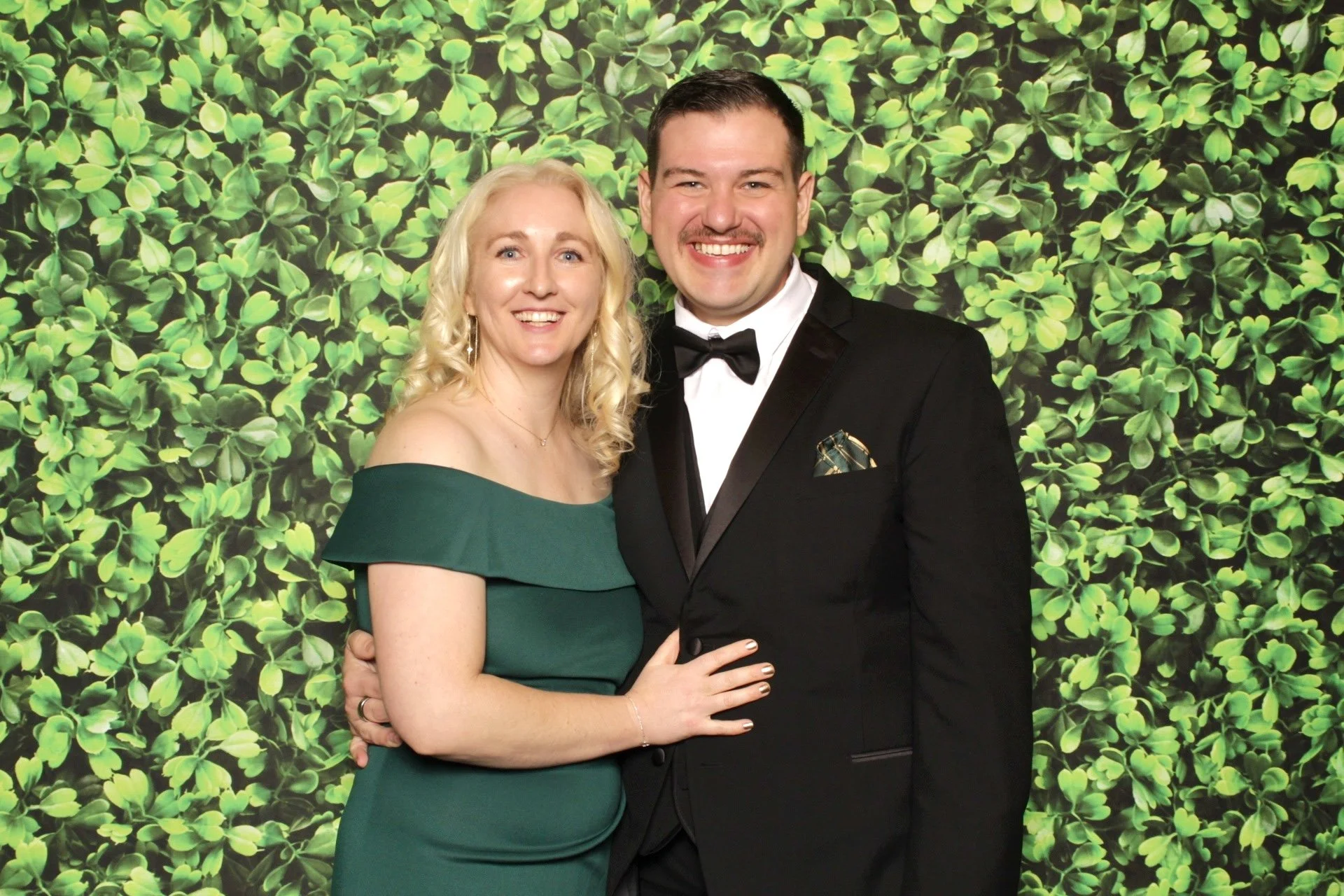 A smiling man in a black tuxedo with a bow tie and a woman in a green off-the-shoulder dress, standing in front of a leafy green backdrop.