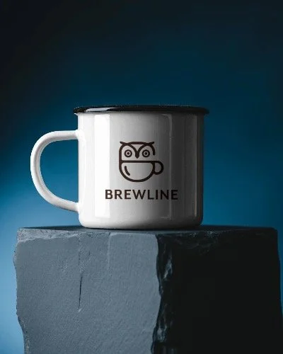 A white mug with a black lid and a cartoon owl face logo, labeled 'BREWLINE,' placed on a dark gray stone block against a dark background.