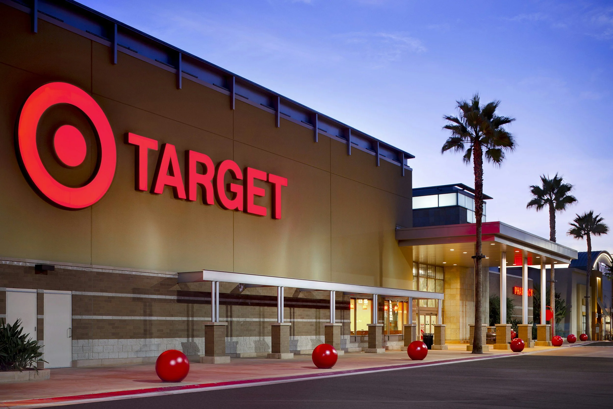 Target store with palm trees outside during dusk, illuminated signage, red spherical decorations along sidewalk, pharmacy sign visible.
