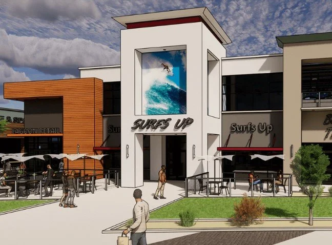 A modern shopping center with a surfing theme, featuring a large mural of a surfer riding a wave on the front facade labeled "Surfs Up." There are outdoor seating areas with umbrellas, trees, and people walking around.