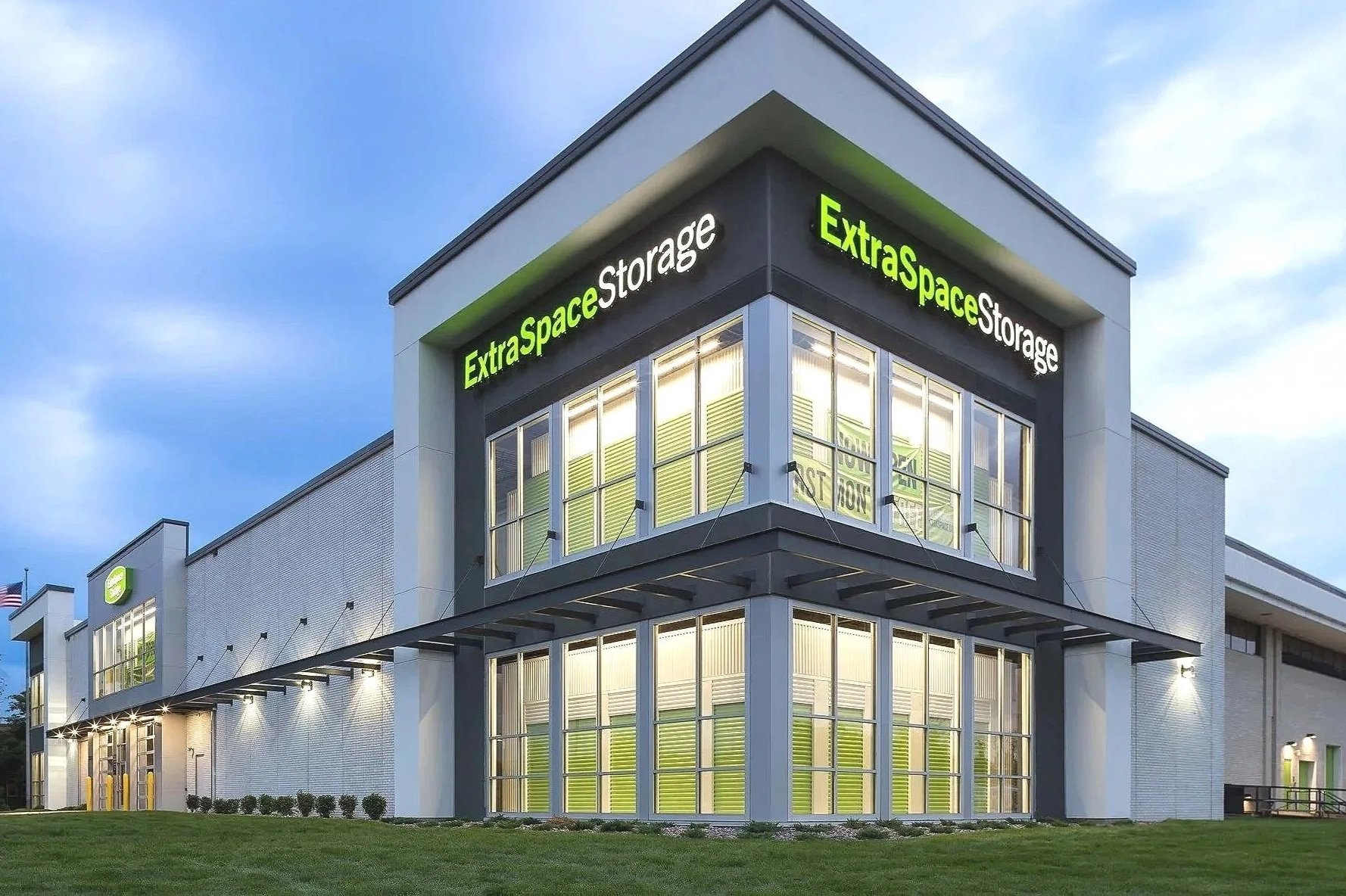 Exterior view of Extra Space Self Storage building at dusk, with illuminated signage and large windows, featuring a manicured lawn in front.