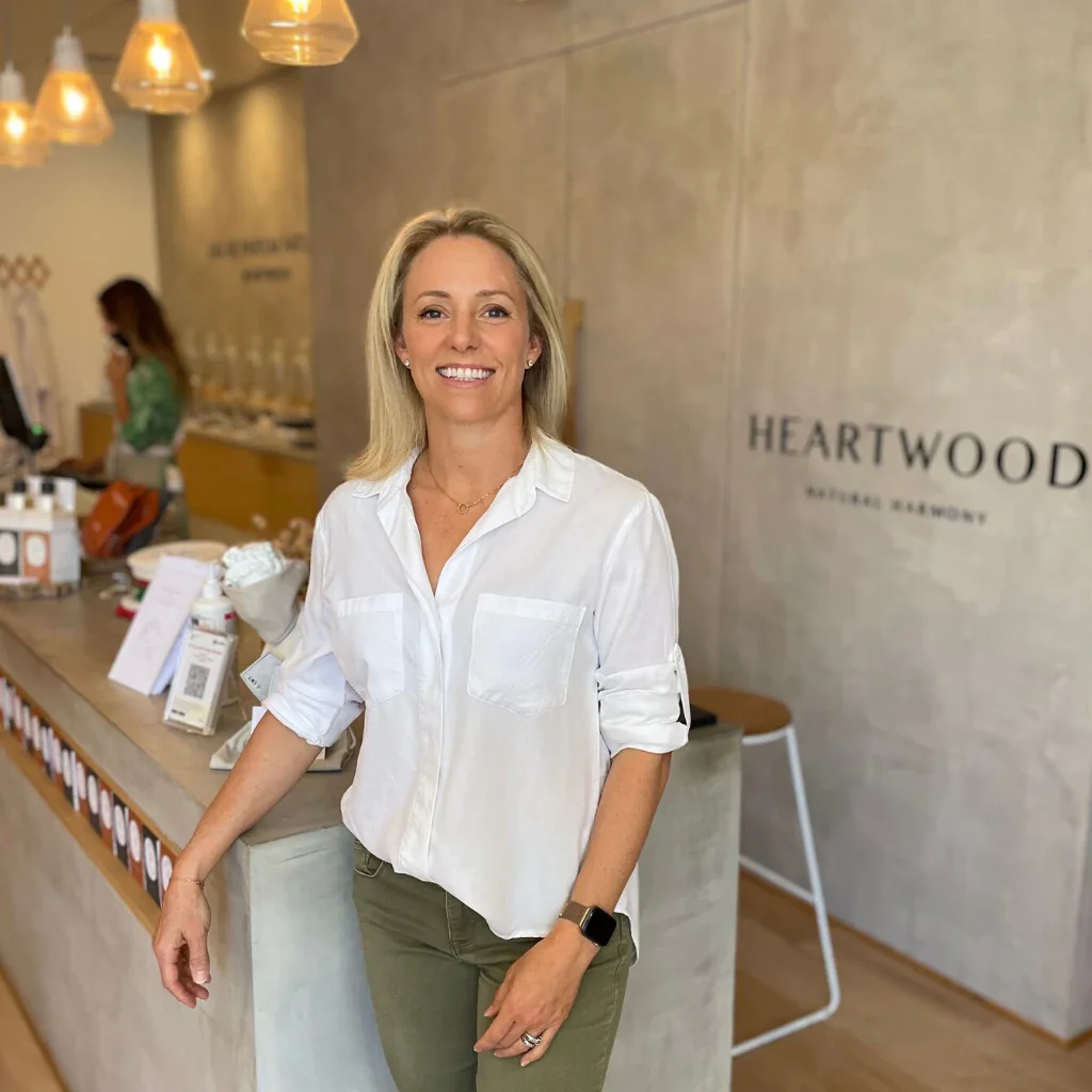 A woman with blonde hair smiling and wearing a white shirt and green pants standing at a reception desk in a hotel lobby.