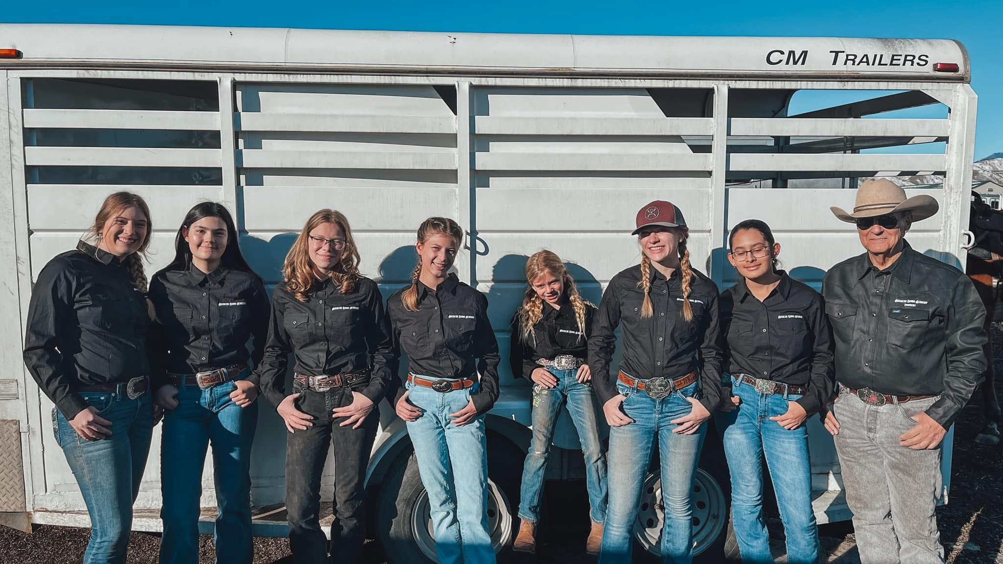 Group of eight young women and one older man dressed in western-style cowboy attire, standing in front of a white trailer with the words "CM Trailers" on it, outdoors on a sunny day.