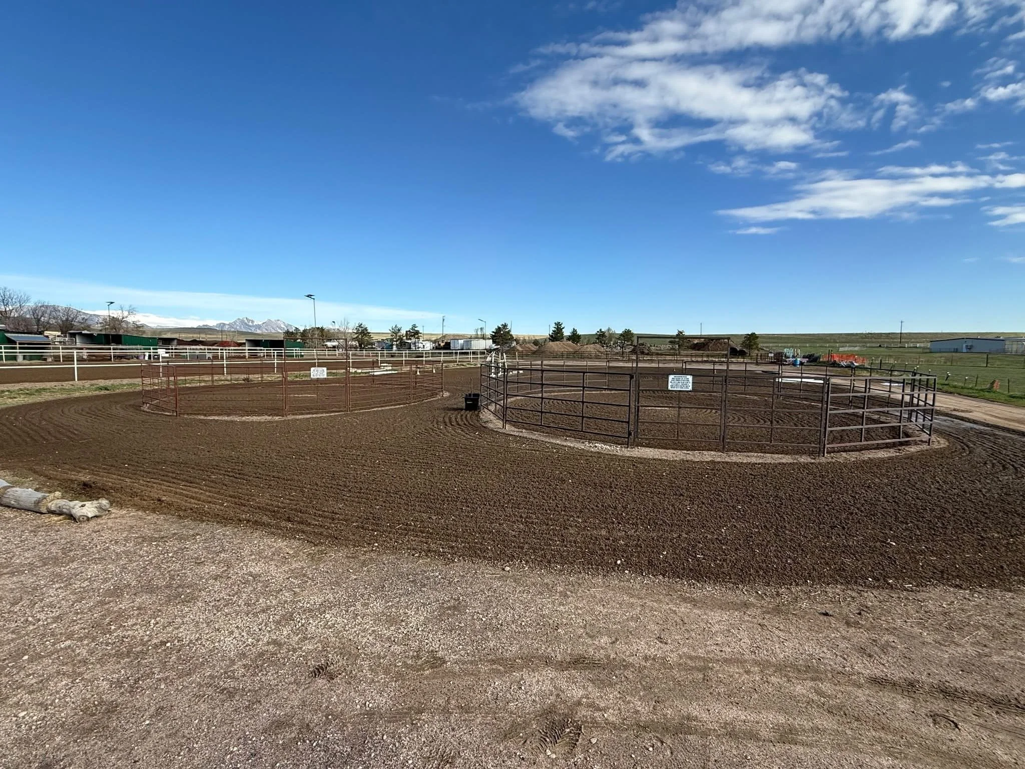 Empty horse riding arenas with dirt footing, surrounded by metal fencing, under a bright blue sky with scattered clouds and mountain range in the background.