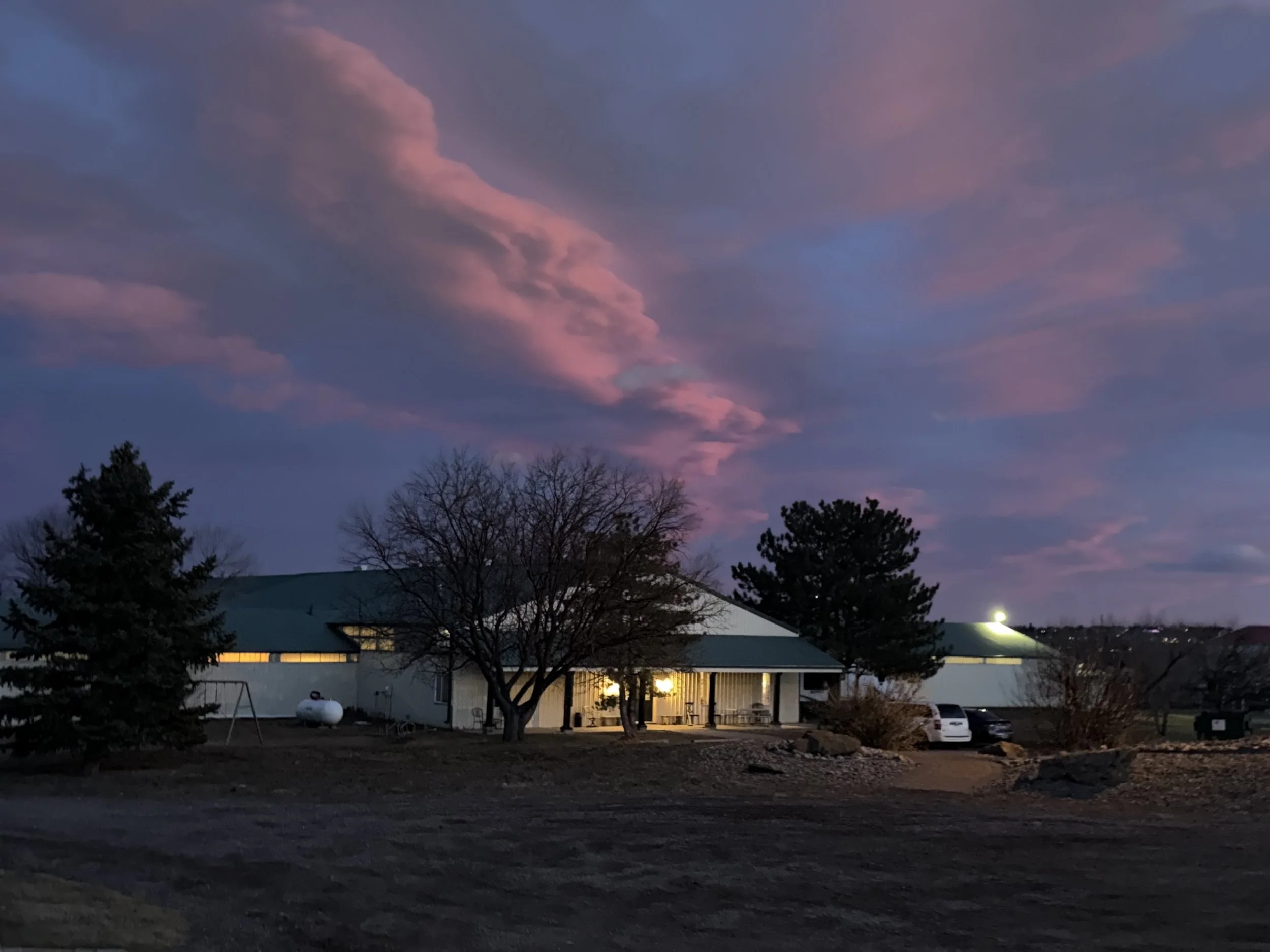A rural scene during twilight featuring a building with a green roof and white siding, several trees, parked cars, and a sky with pink and purple clouds.