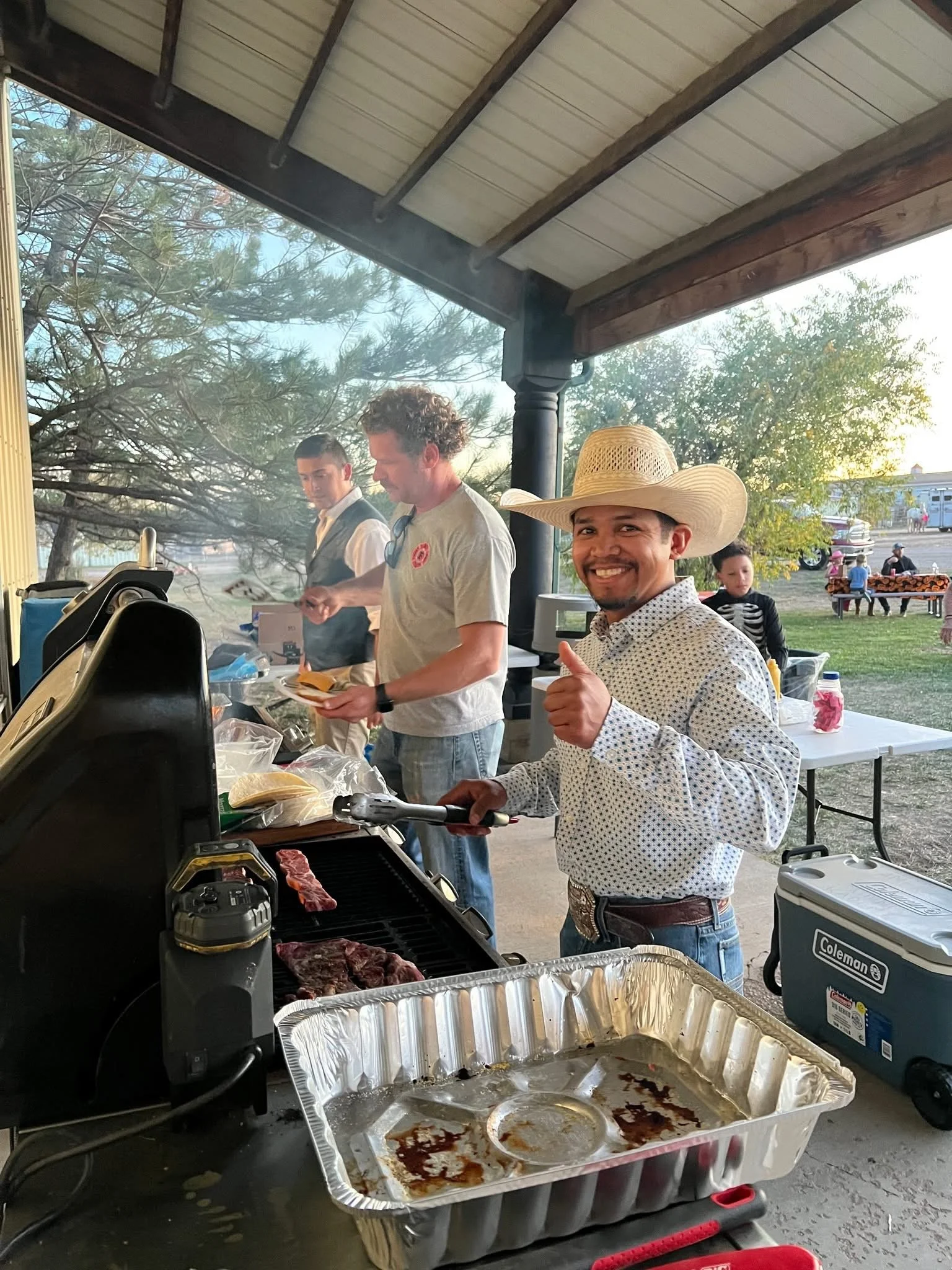 Smiling man in a cowboy hat giving a thumbs up while grilling meat at an outdoor barbecue.