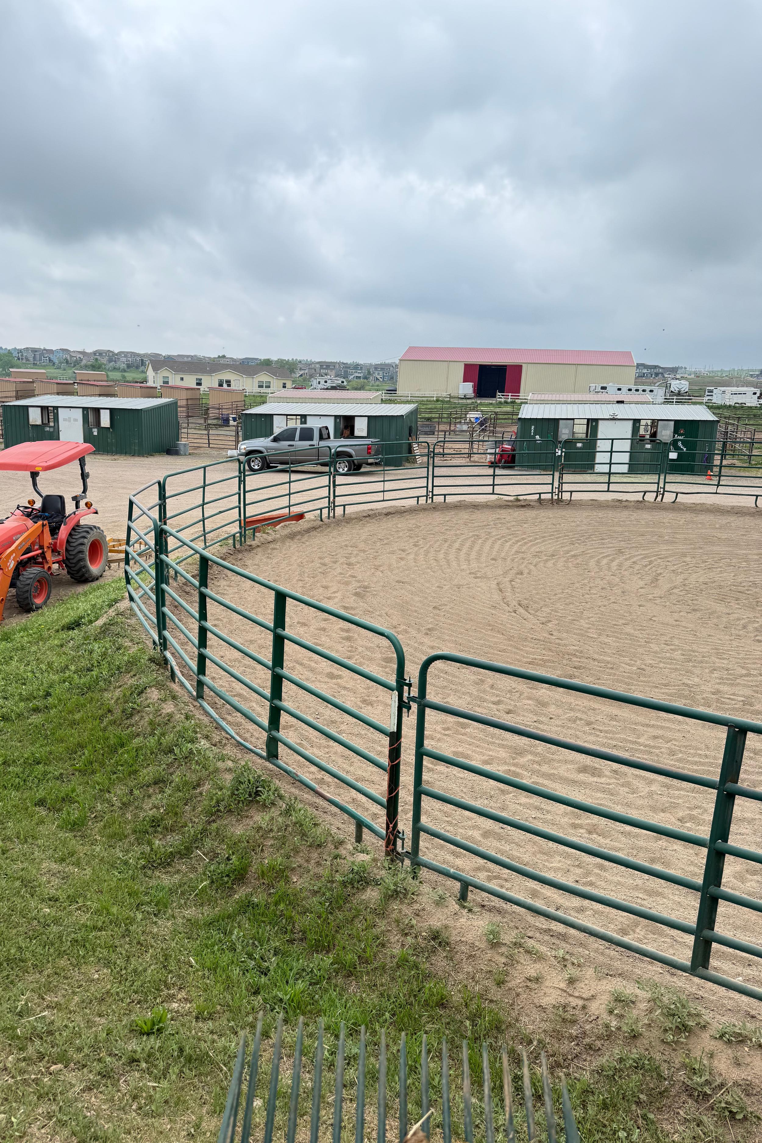 A fenced outdoor rodeo arena with sandy ground, a red tractor on the left, and green metal stalls in the background under a cloudy sky.