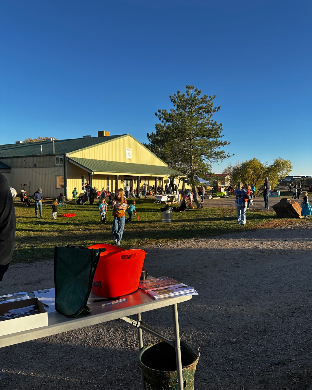 Outdoor community event with people gathered near a yellow building, children playing, tables set up, and a clear blue sky.