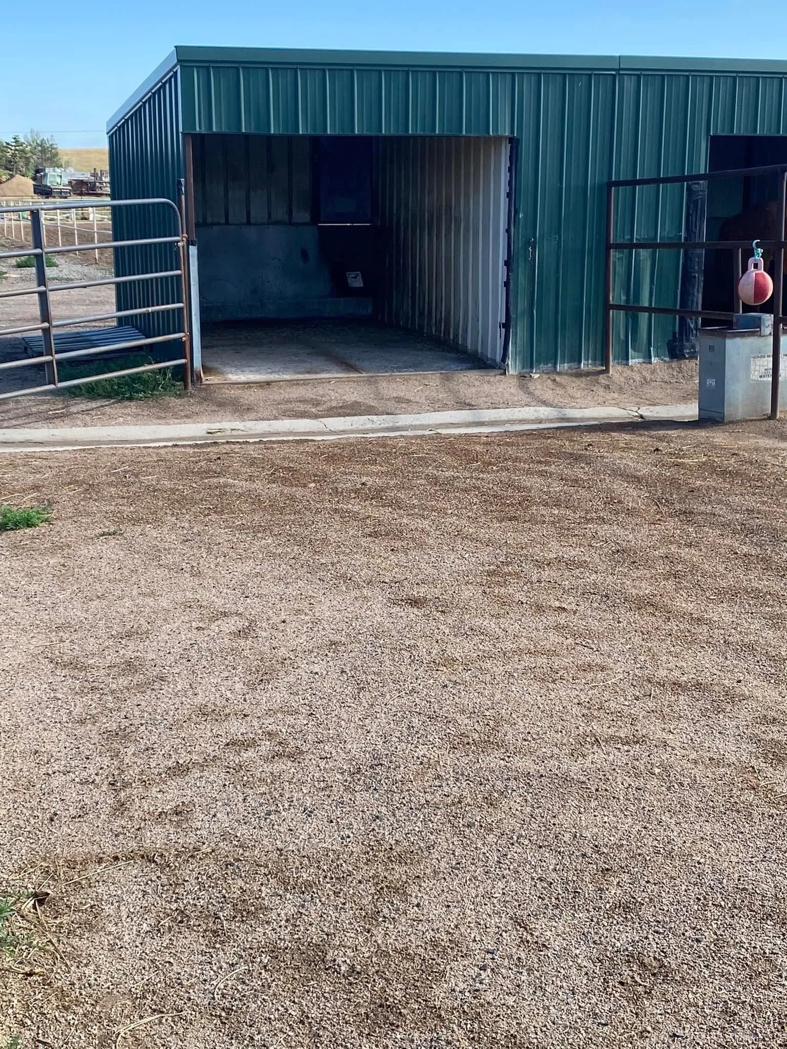 Green metal barn with a wide entry, situated in an outdoor farm or ranch setting, with a gravel ground area in foreground and a small metal railing on either side of the entrance.