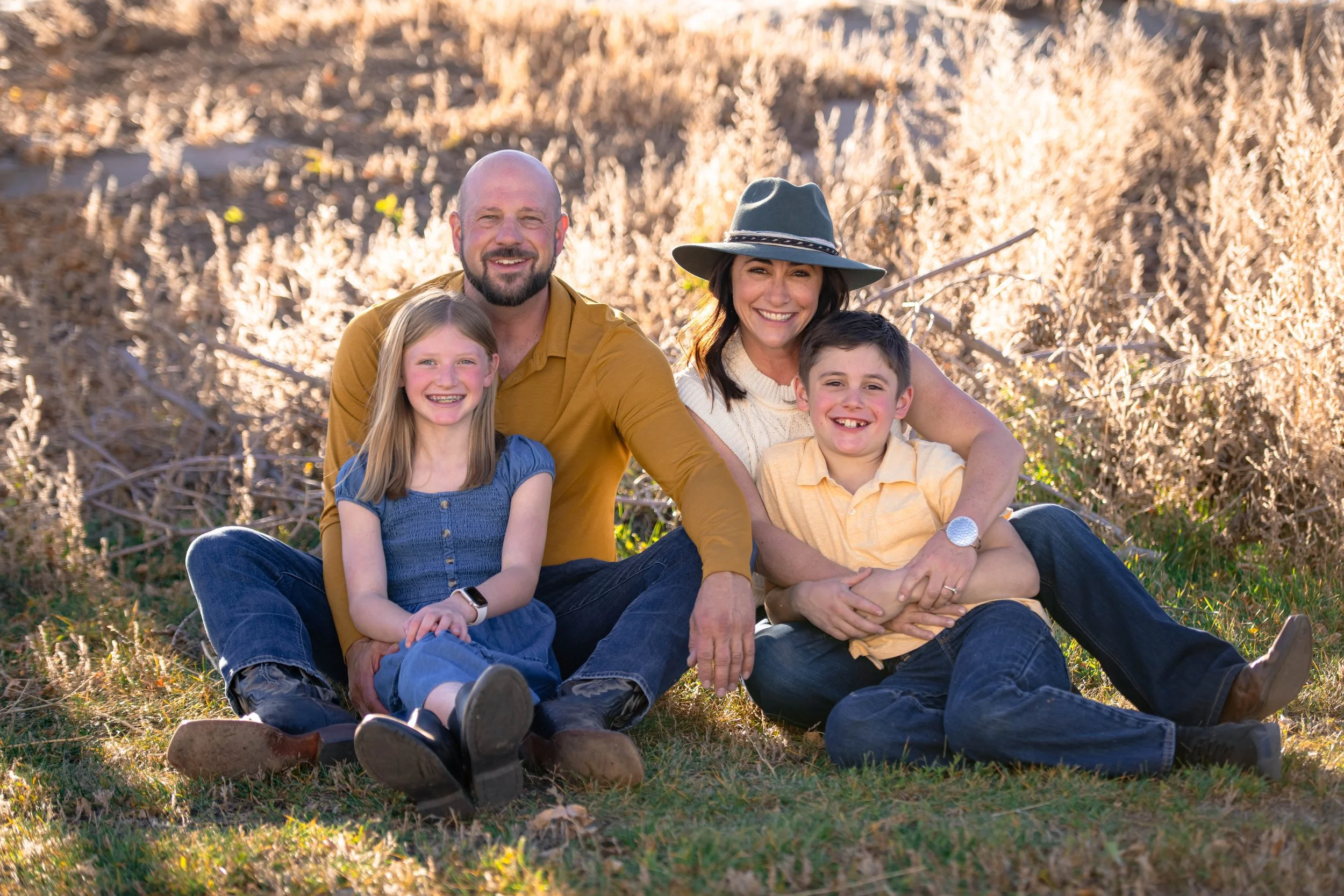 Family of four sitting on the grass outdoors in front of fall foliage, smiling at the camera. The father has a beard and is wearing a mustard yellow shirt, the mother is wearing a wide-brimmed hat and a cream sweater, the daughter has long blonde hair and a blue dress, and the son is wearing a yellow shirt.